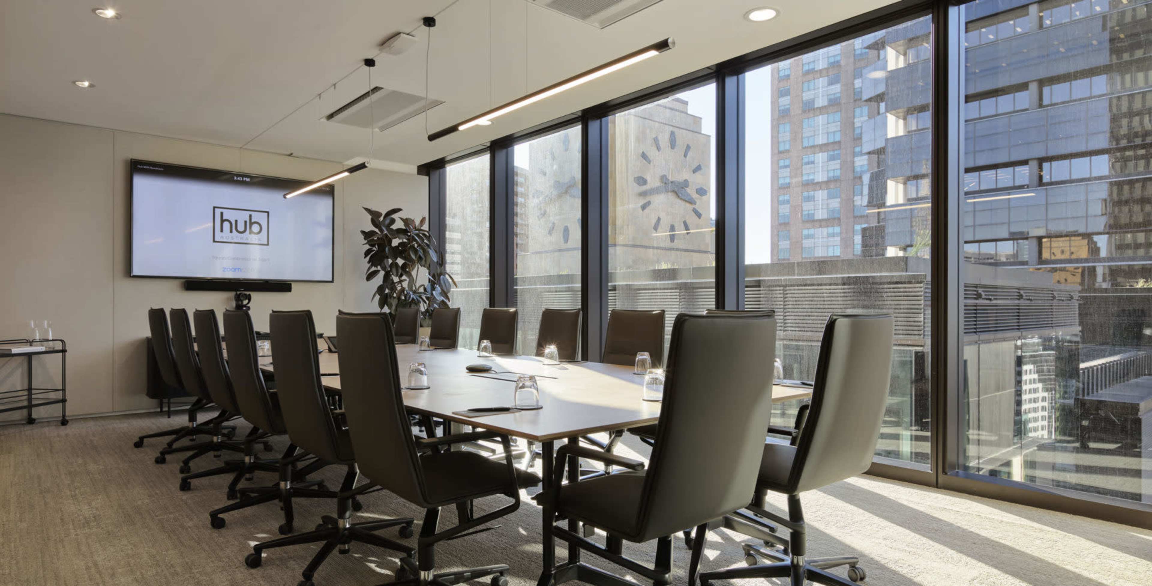 A modern conference room features a long table with black ergonomic chairs, large windows revealing a cityscape, and a wall-mounted screen displaying the logo of "hub."