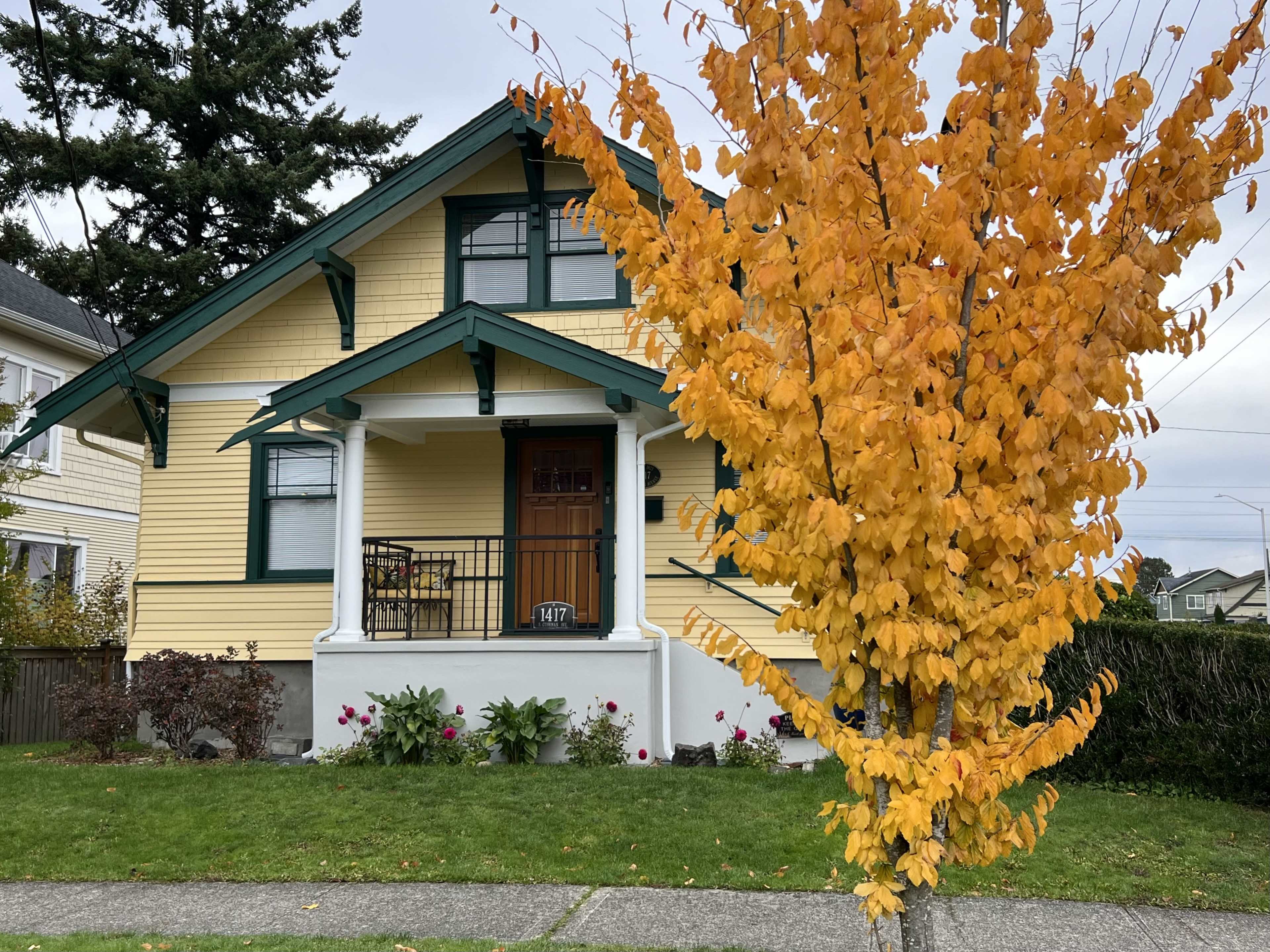 A yellow house with green trim and a covered porch is paired with a bright orange tree in a residential neighborhood.