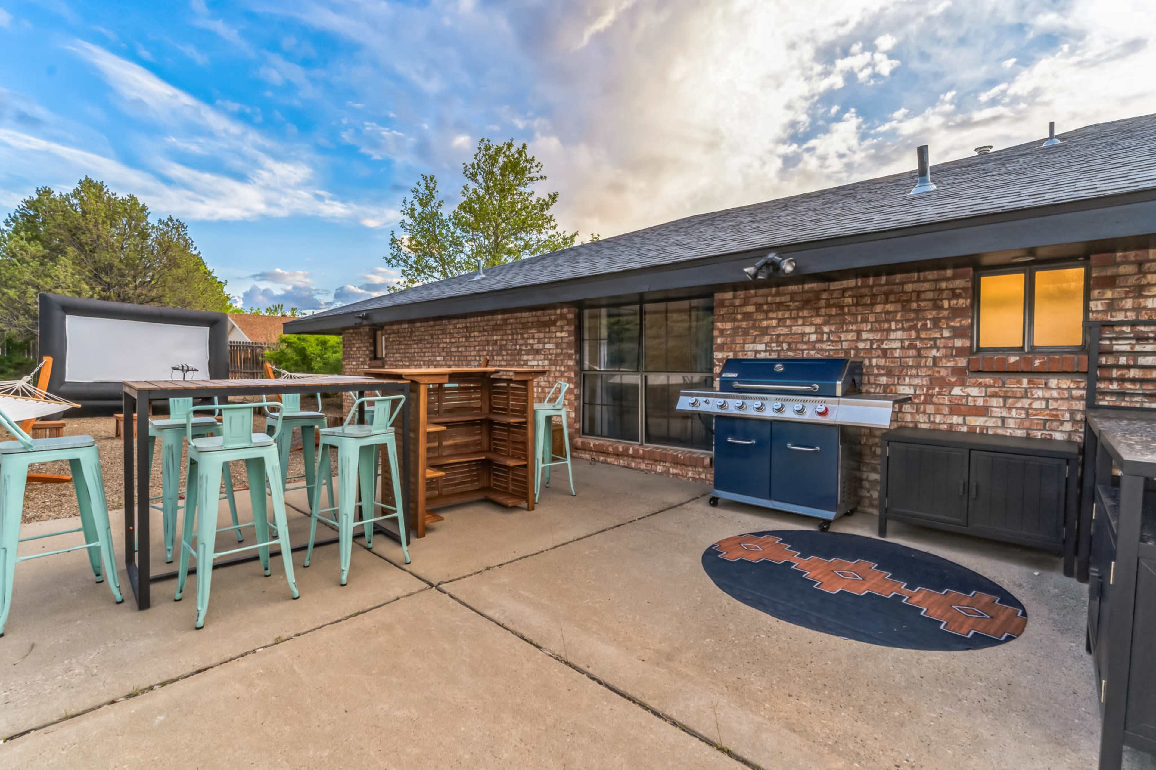 A patio area featuring a blue grill, high-top tables with stools, and a backdrop of trees and clouds.