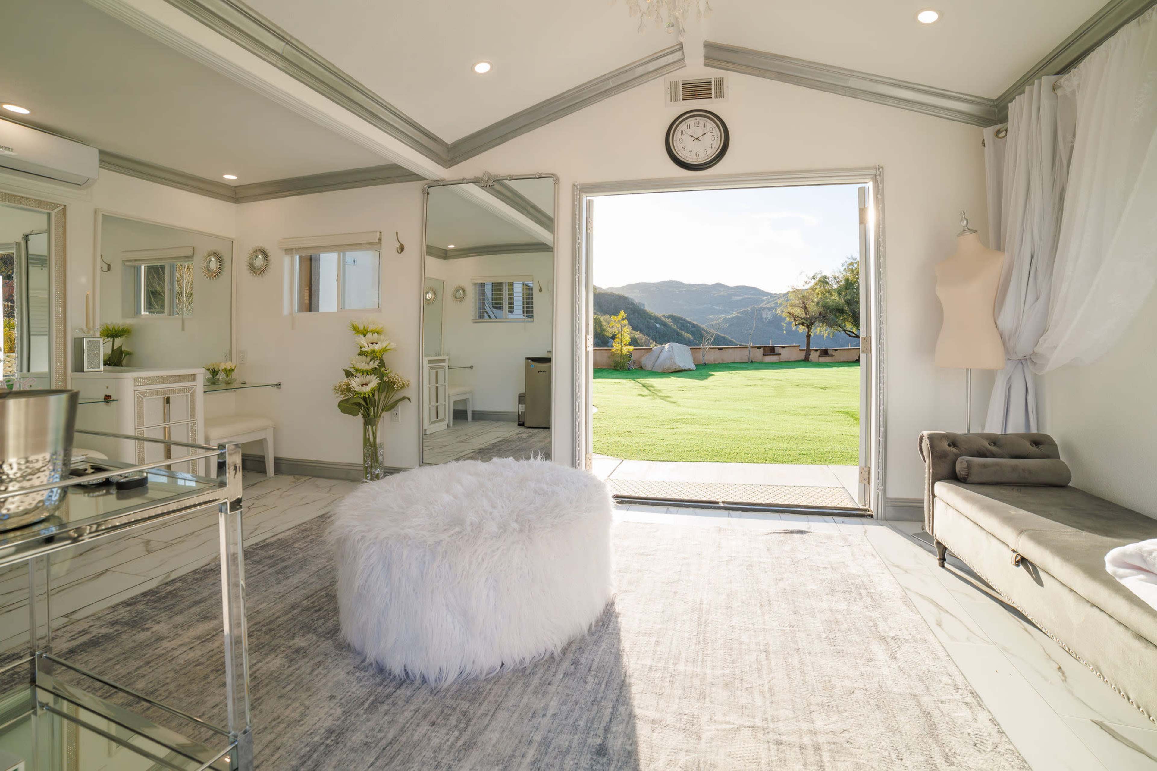 A spacious room with large windows opens to a green outdoor area, featuring a round white fur pouf, a gray chaise lounge, and elegant decor.