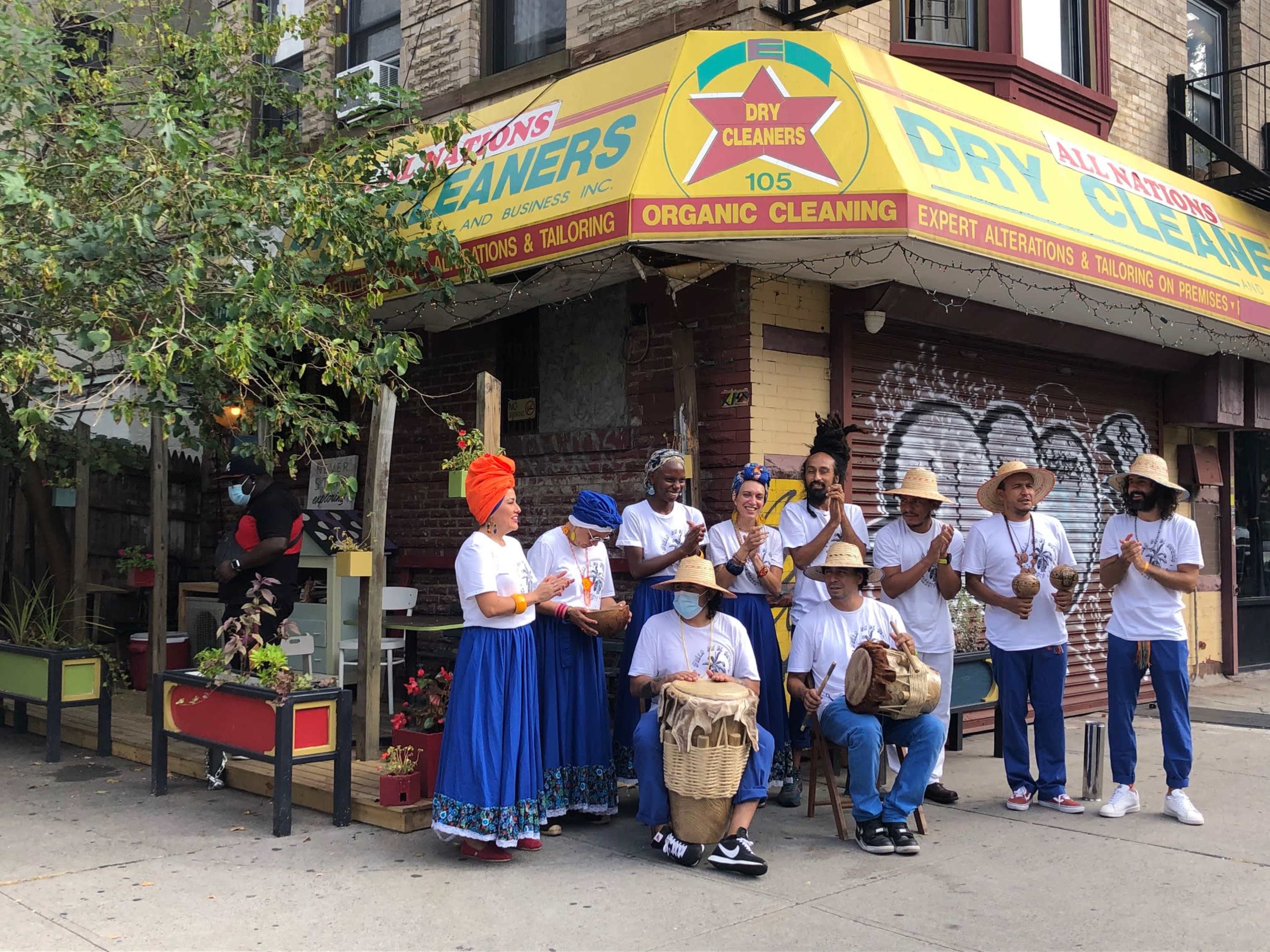 A group of people in traditional attire stand outside a dry cleaning shop, gathered around a woman holding a basket, while others clap and play instruments.