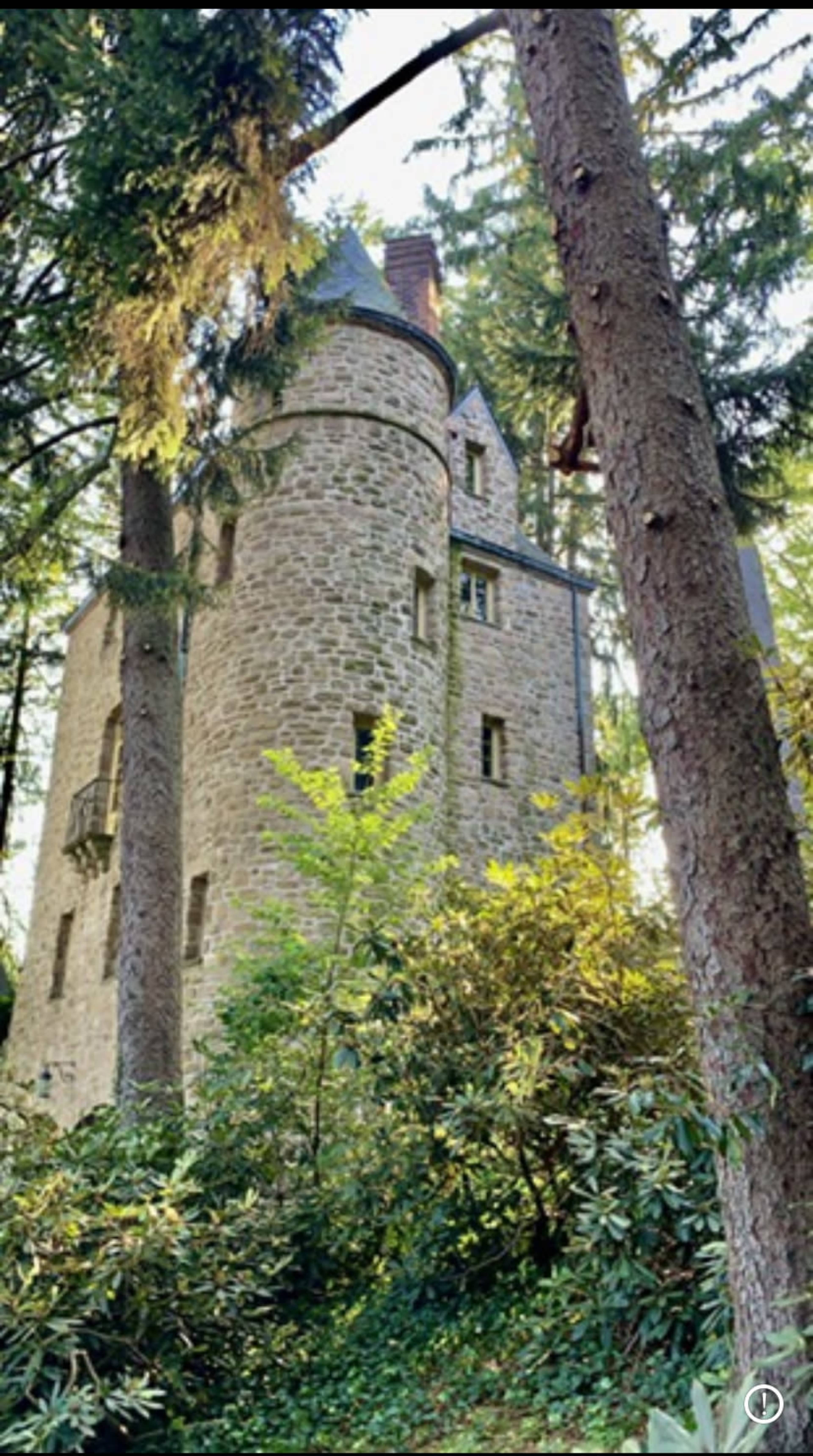 A stone tower with several windows and a balcony is nestled among tall trees and dense foliage.