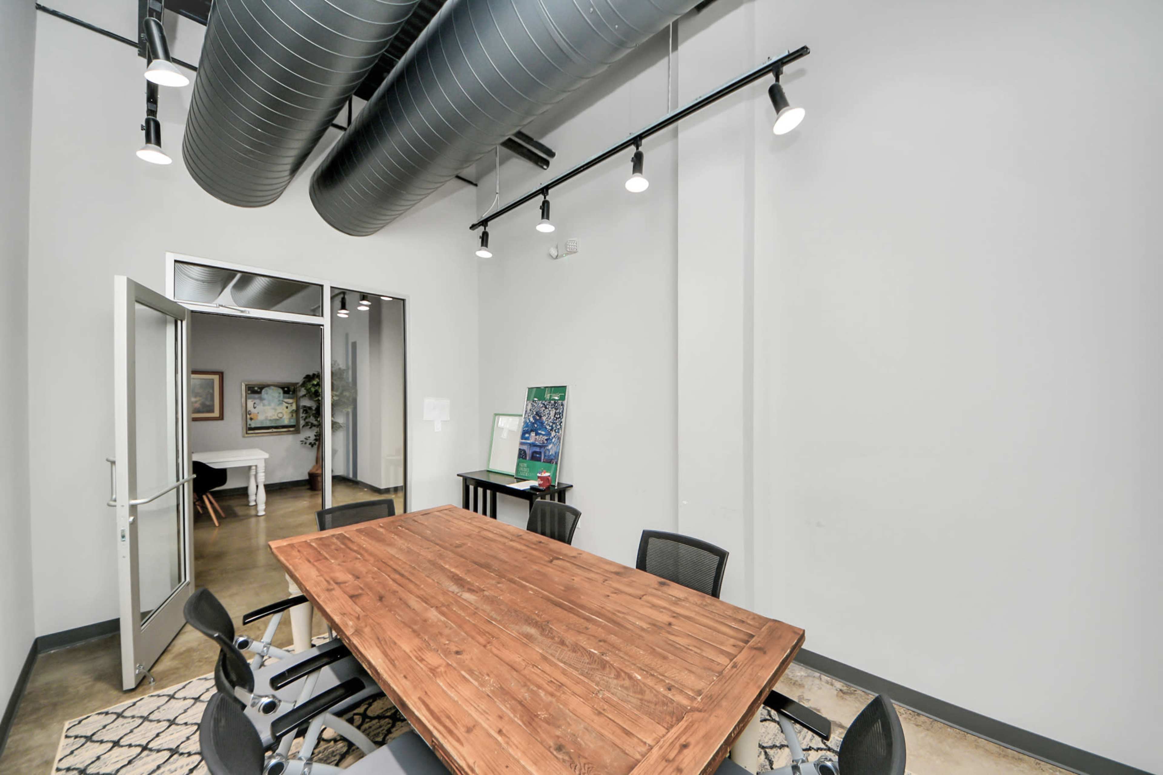 The image shows a modern conference room featuring a large wooden table surrounded by black chairs, with exposed ductwork and overhead lighting.