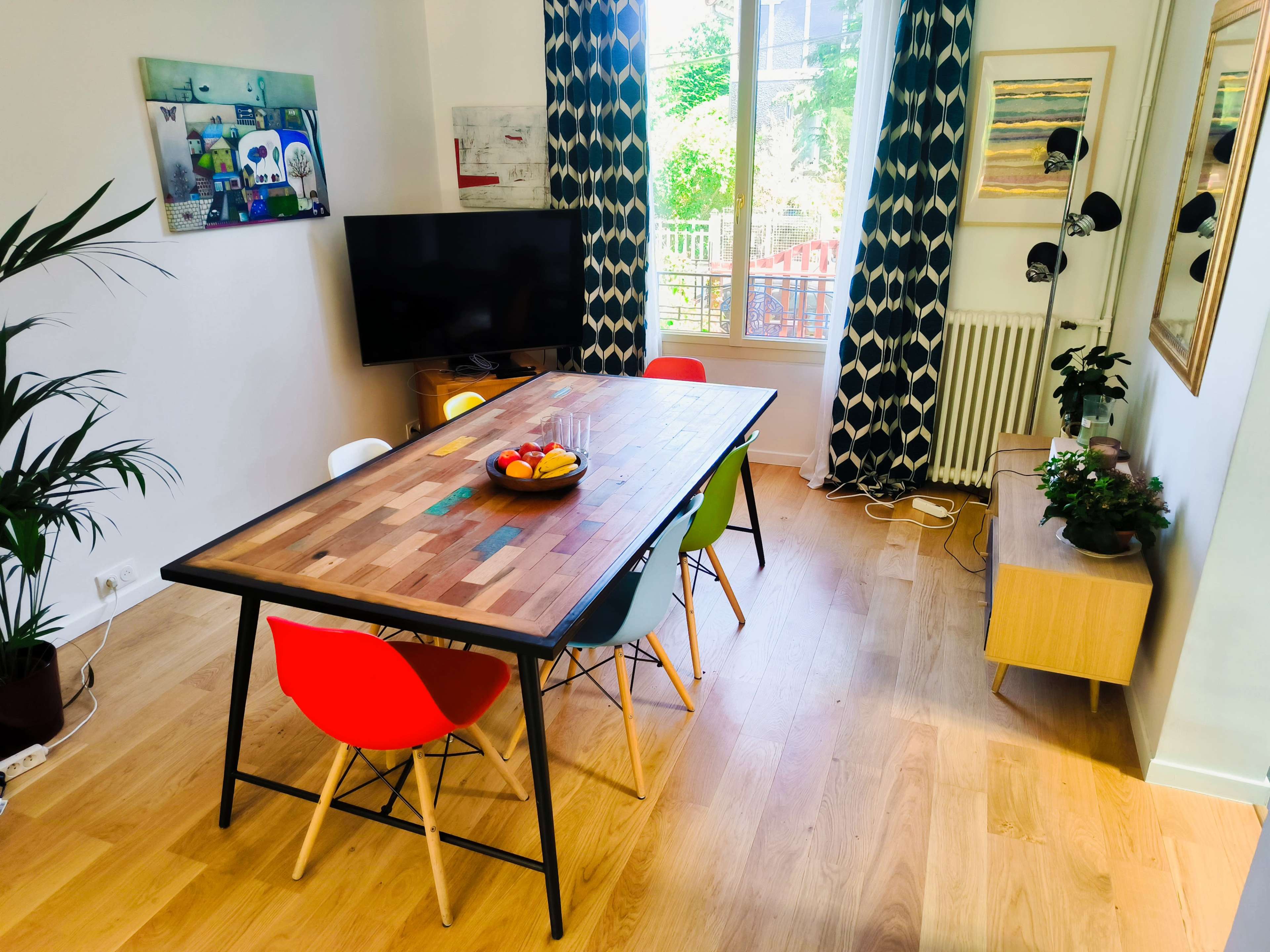 A dining area features a wooden table surrounded by colorful chairs, a television on the wall, and large windows allowing natural light to enter.
