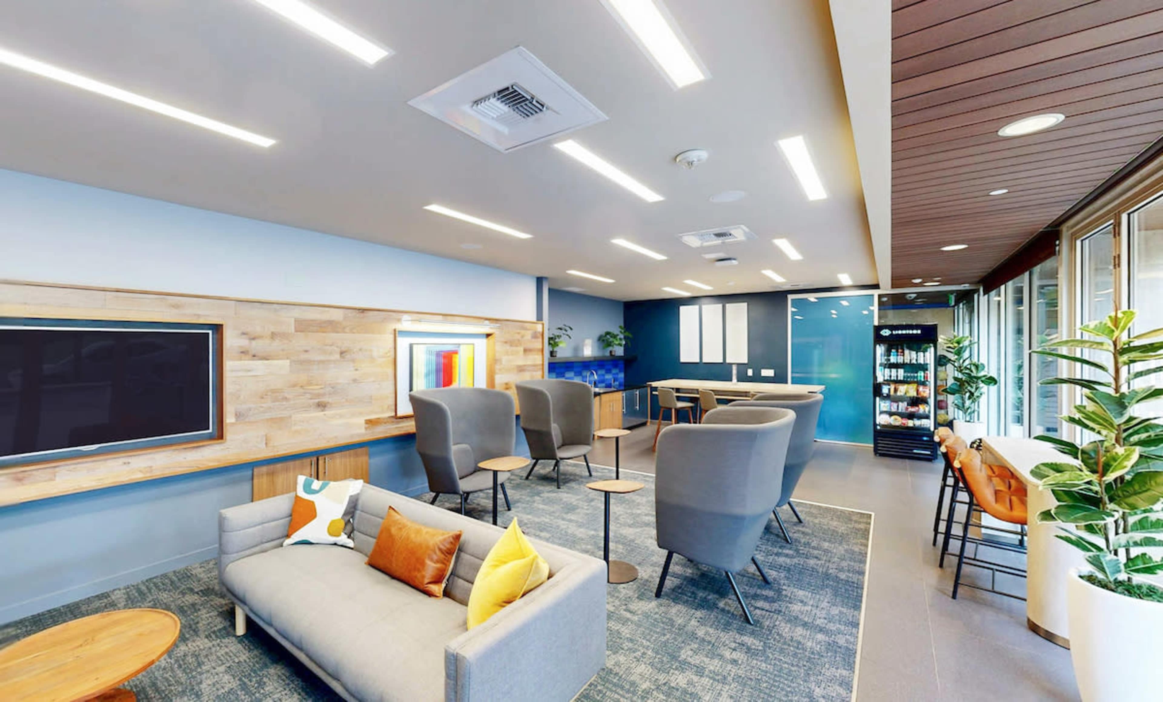 The image shows a modern lounge area featuring a gray sofa, several armchairs, a coffee table, and a wall-mounted television in a well-lit space with blue and wooden accents.