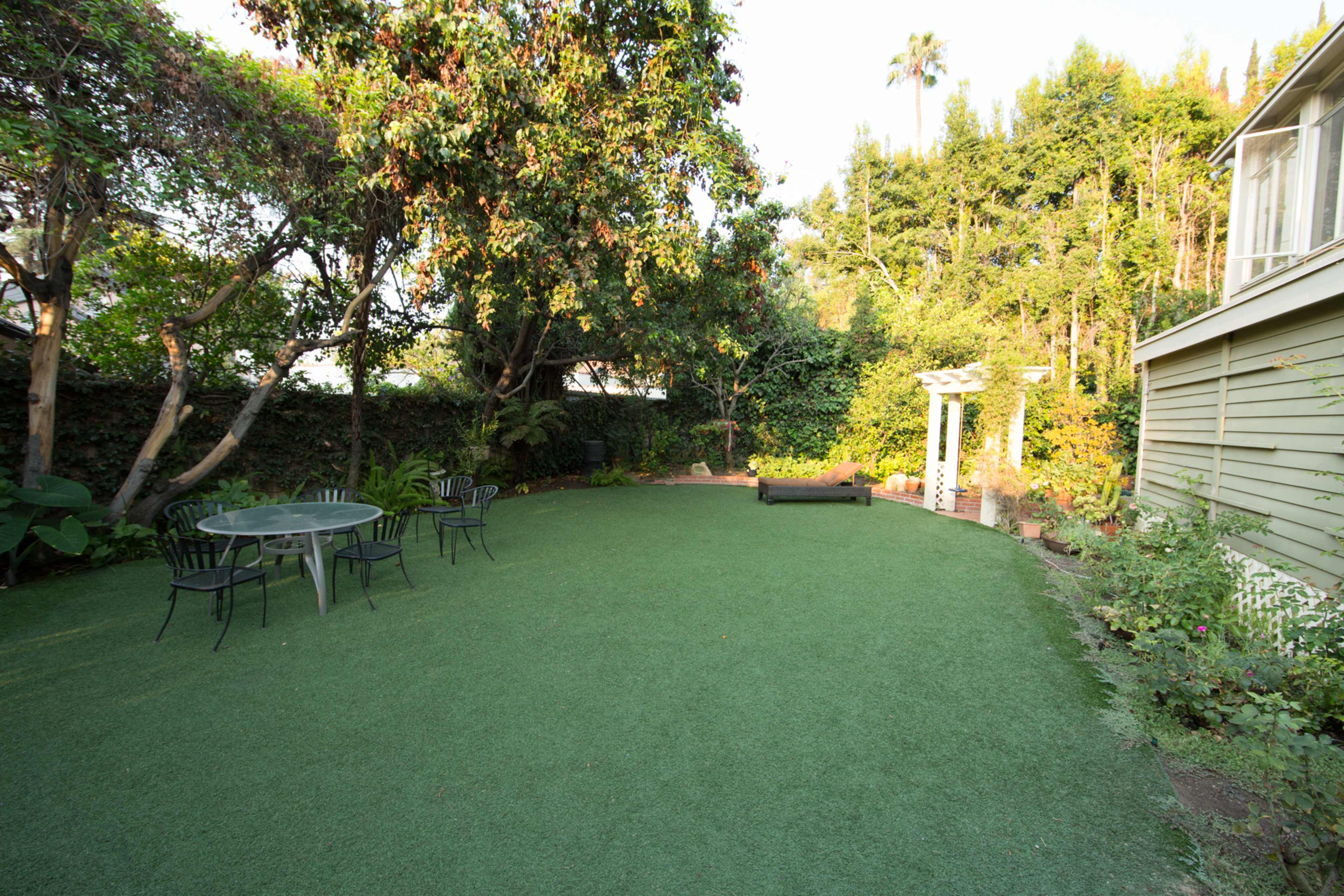 A green, artificial turf lawn with a table and chairs, surrounded by trees and a house on one side.