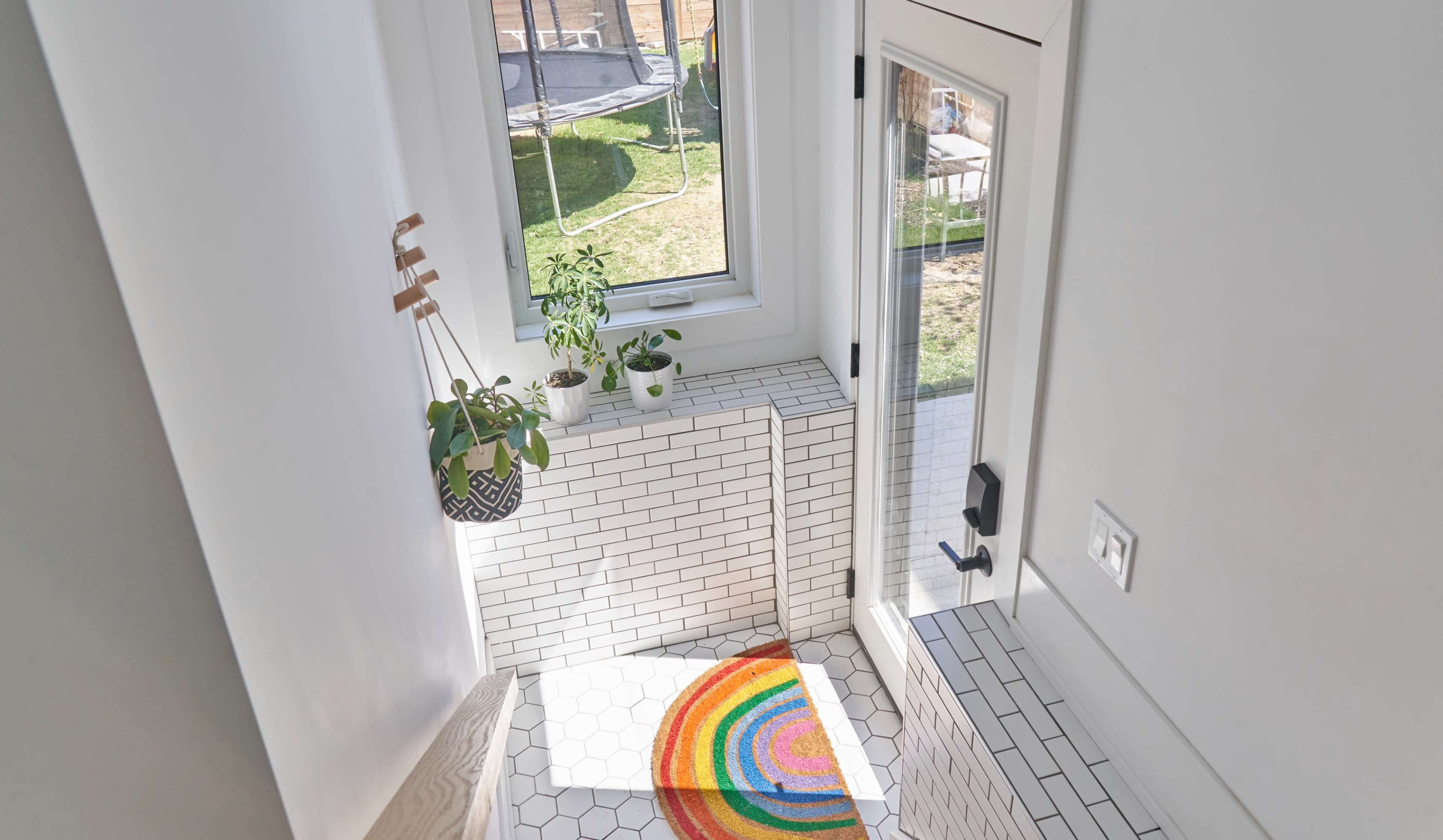 A bright entryway features white tile walls and a colorful striped rug leading to a door with large windows overlooking a grassy area.