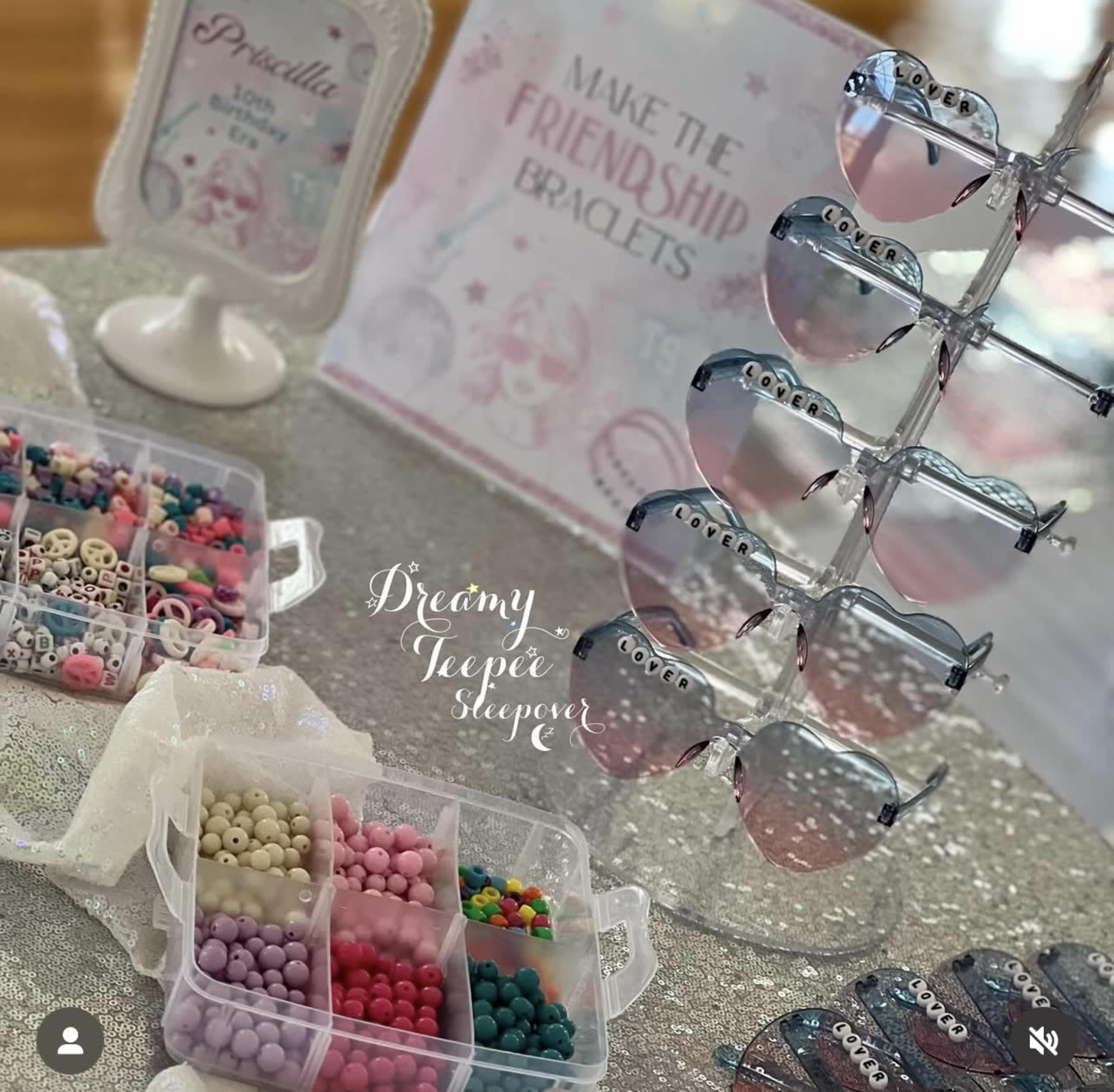 The image shows a table setup featuring a display of sunglasses, a sign about making friendship bracelets, and a container filled with colorful beads.