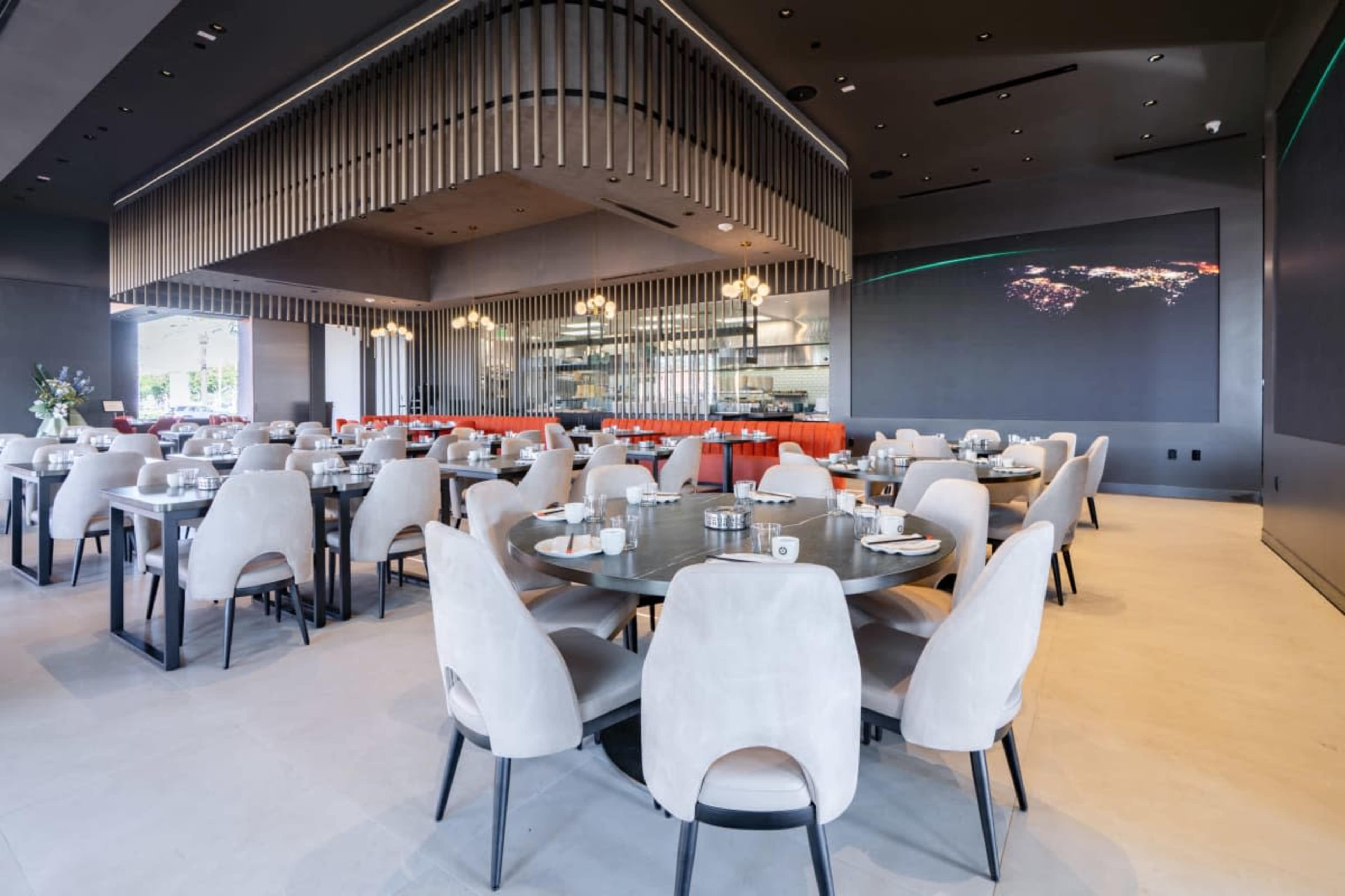 The image shows a modern restaurant interior featuring round tables with chairs, gray walls, and a shimmering bar area in the background.