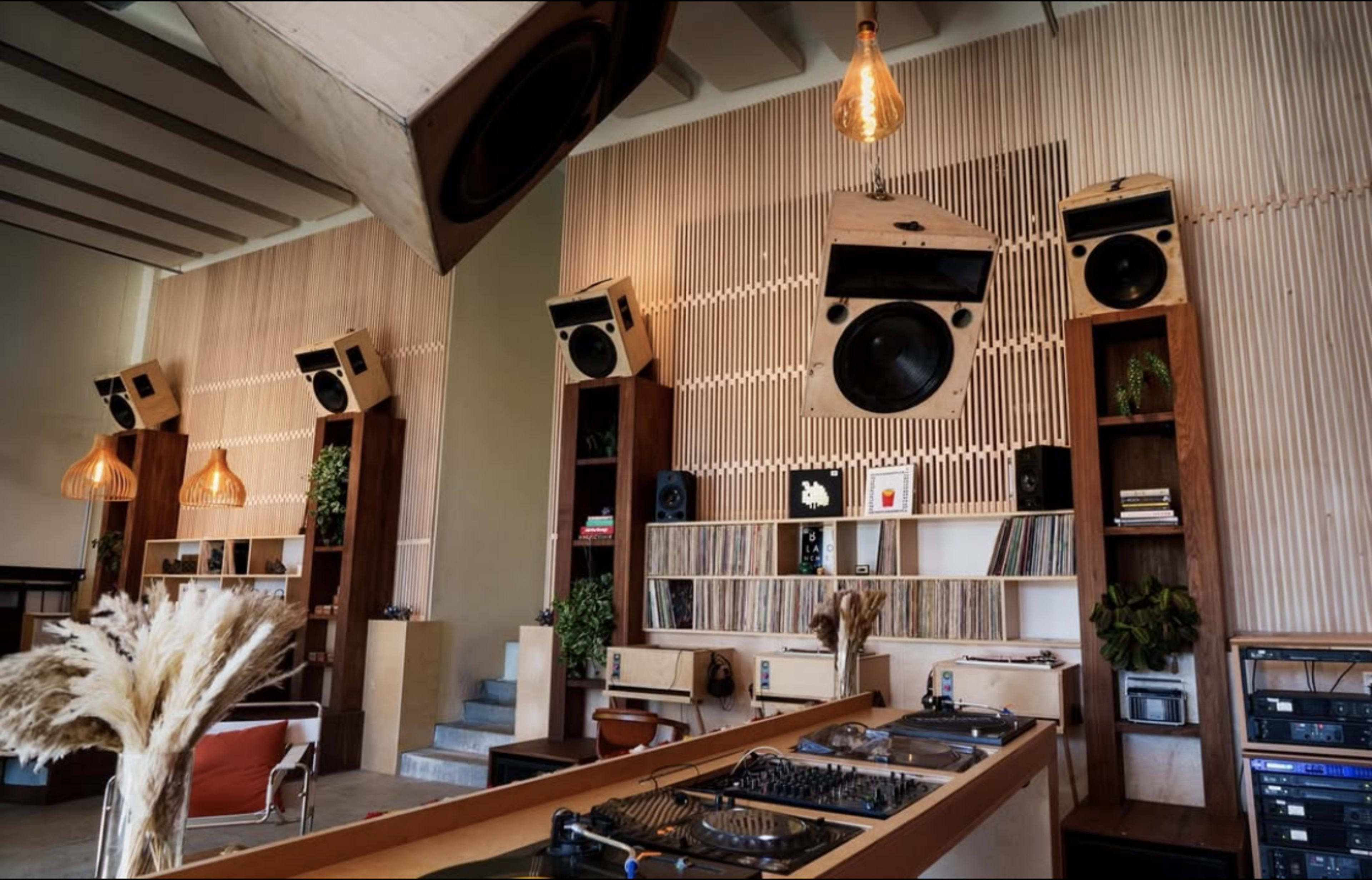 A stylish interior features a wall of vinyl records and large speakers, with a DJ setup in the foreground and warm lighting accents.