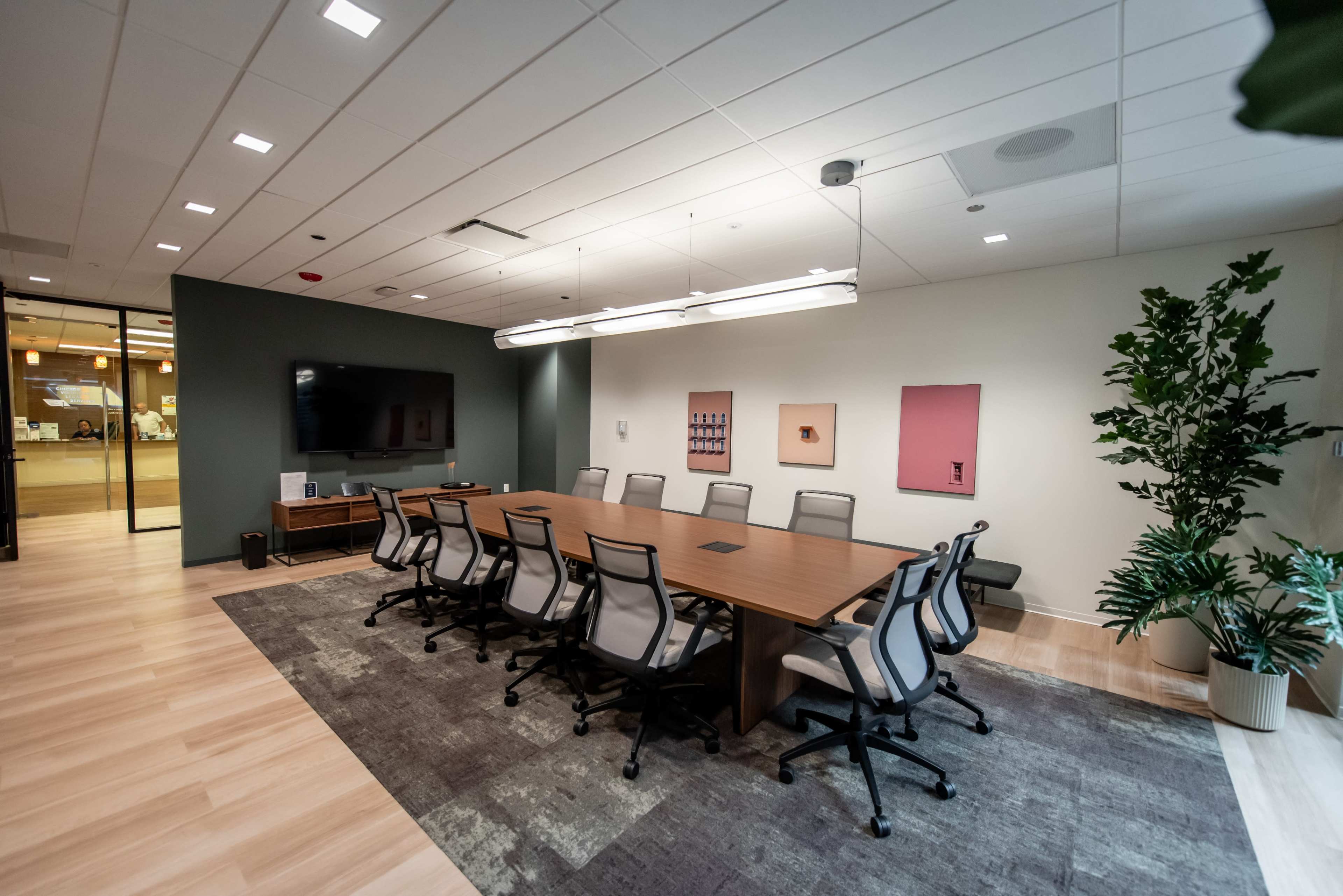 A modern conference room features a large wooden table surrounded by ergonomic chairs, with a display screen mounted on the wall and artwork hanging nearby.
