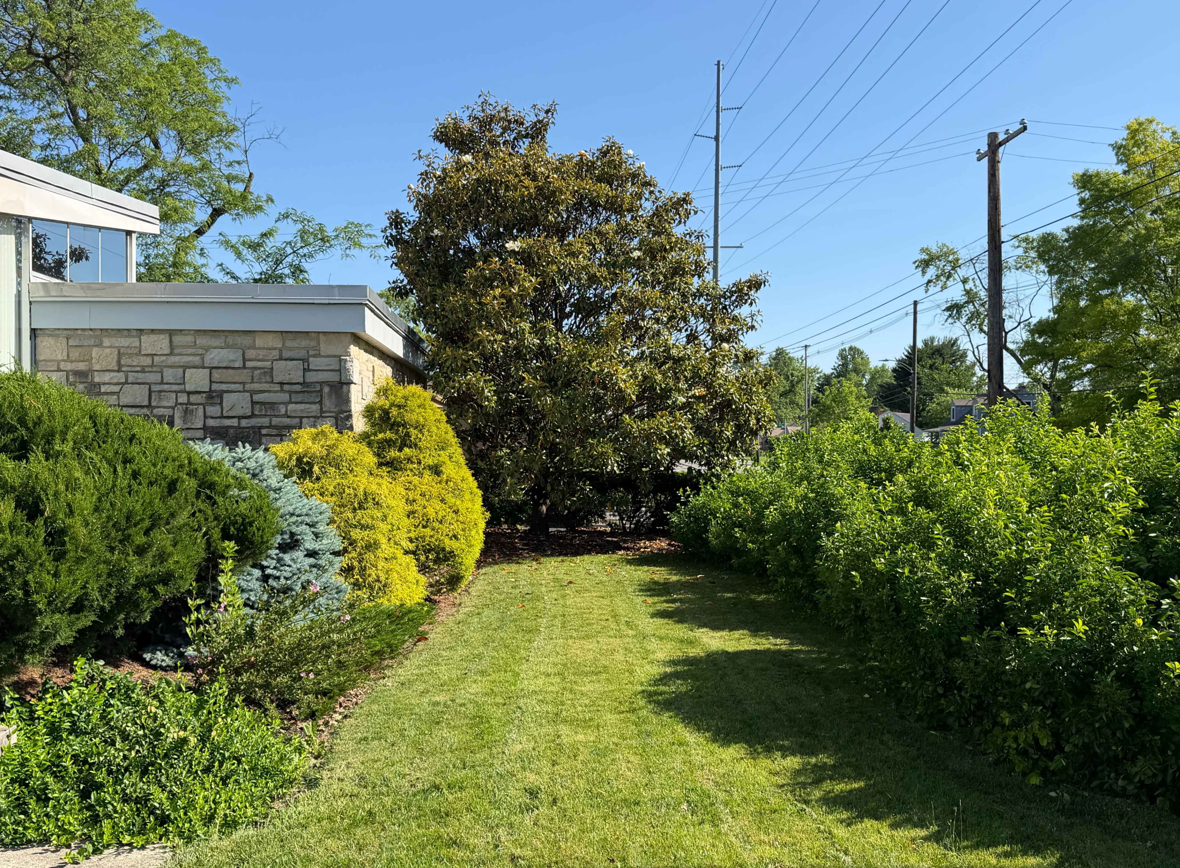 A well-maintained garden path runs alongside a stone building, flanked by various greenery and trees under a clear blue sky.