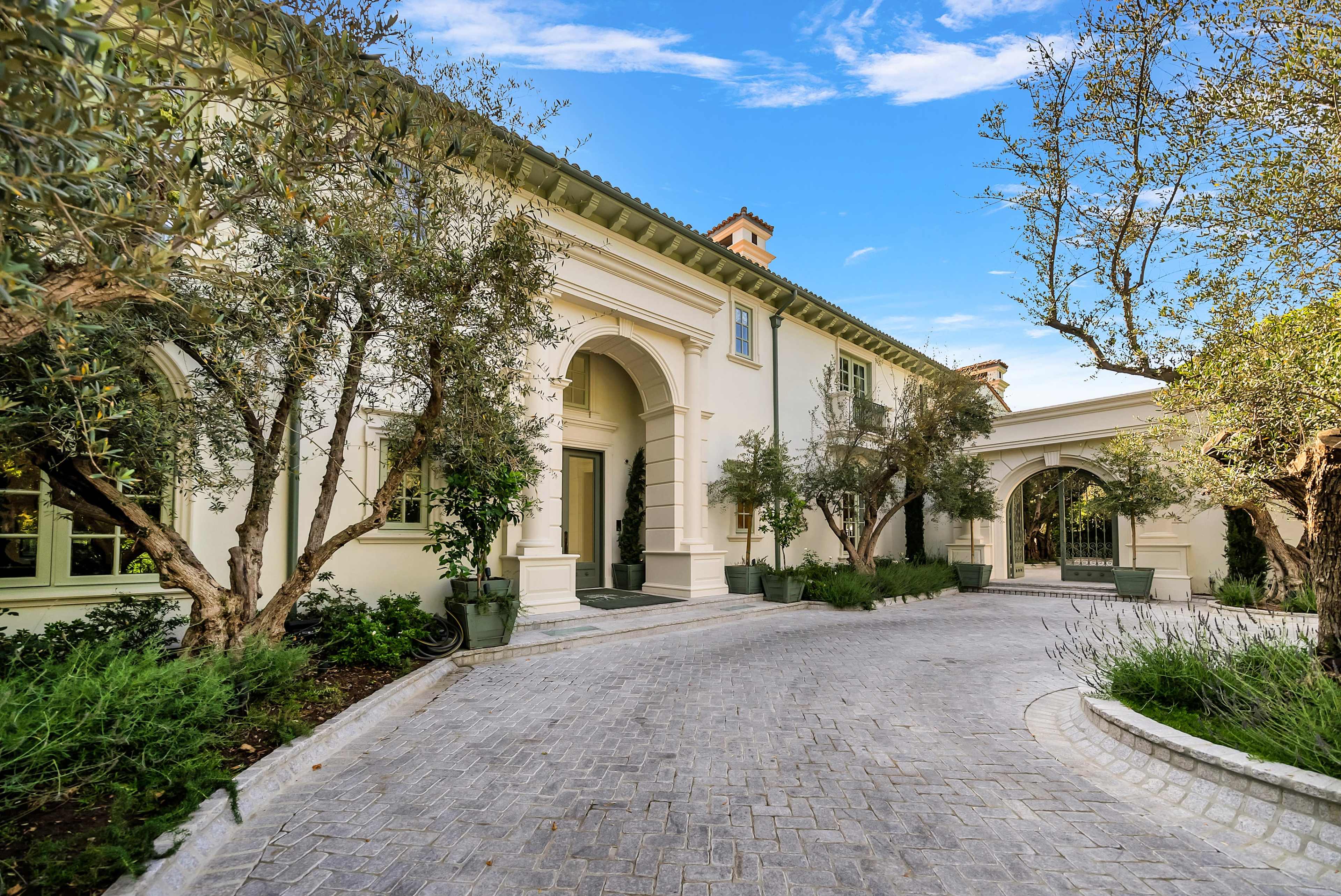 The image shows a luxurious residential exterior with a paved driveway, landscaped olive trees, and an arched entrance to the house.