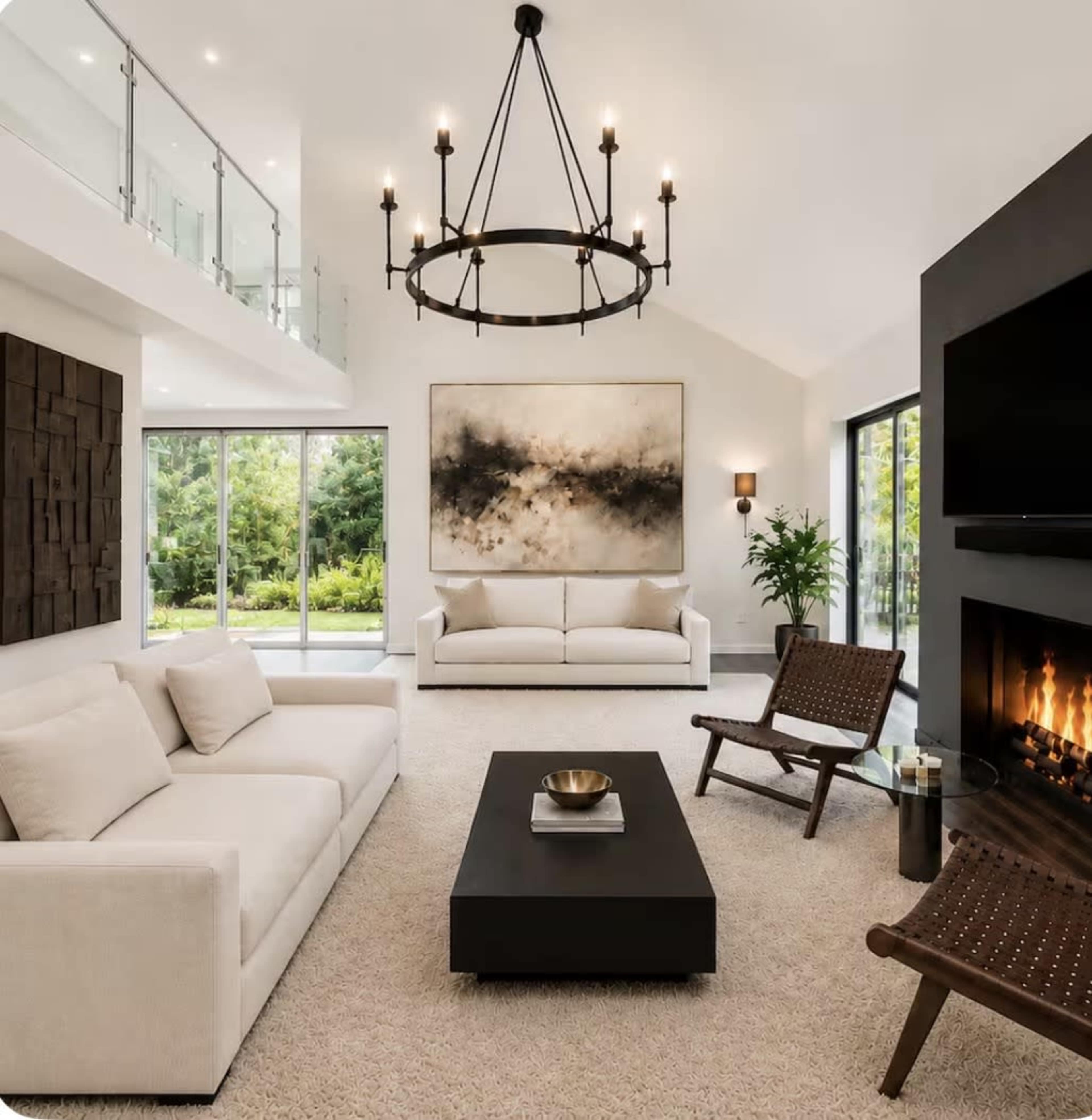 A modern living room features a white sofa and armchairs, a black coffee table, a large artwork, and a fireplace, surrounded by large windows that overlook greenery.