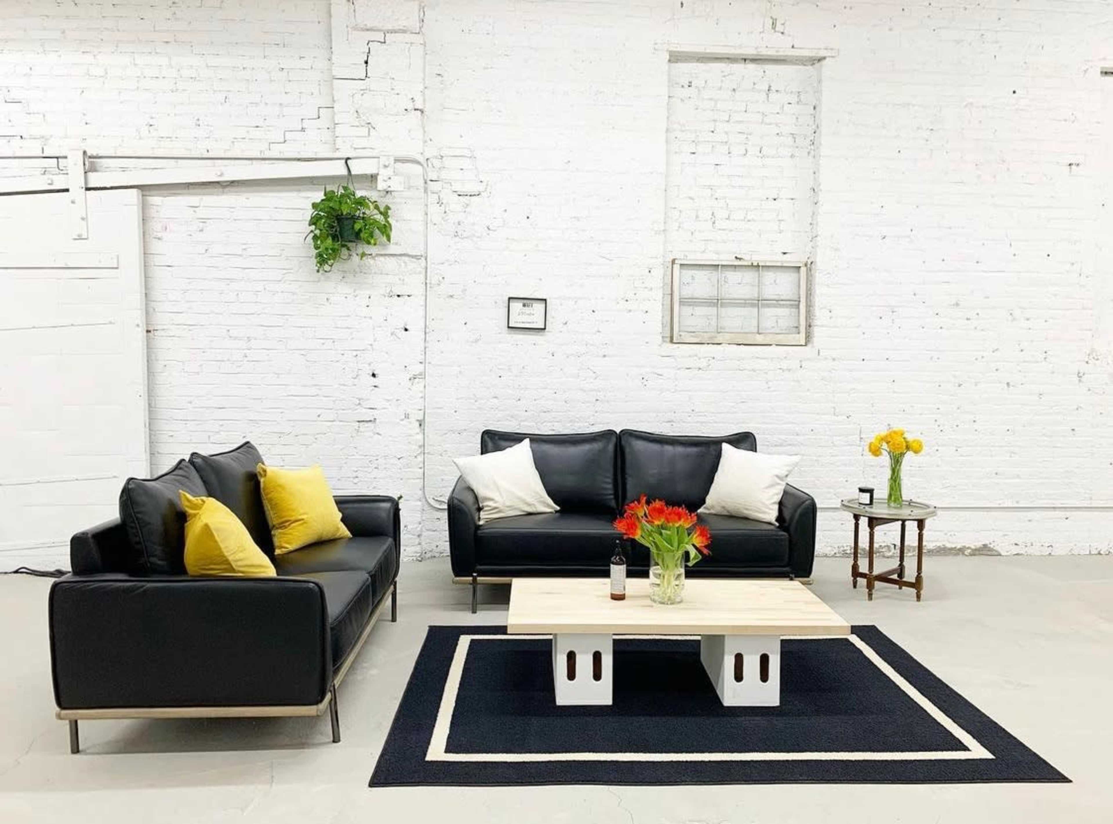 A minimalist living area features two black sofas with yellow pillows, a wooden coffee table, and a small side table set against a white brick wall.