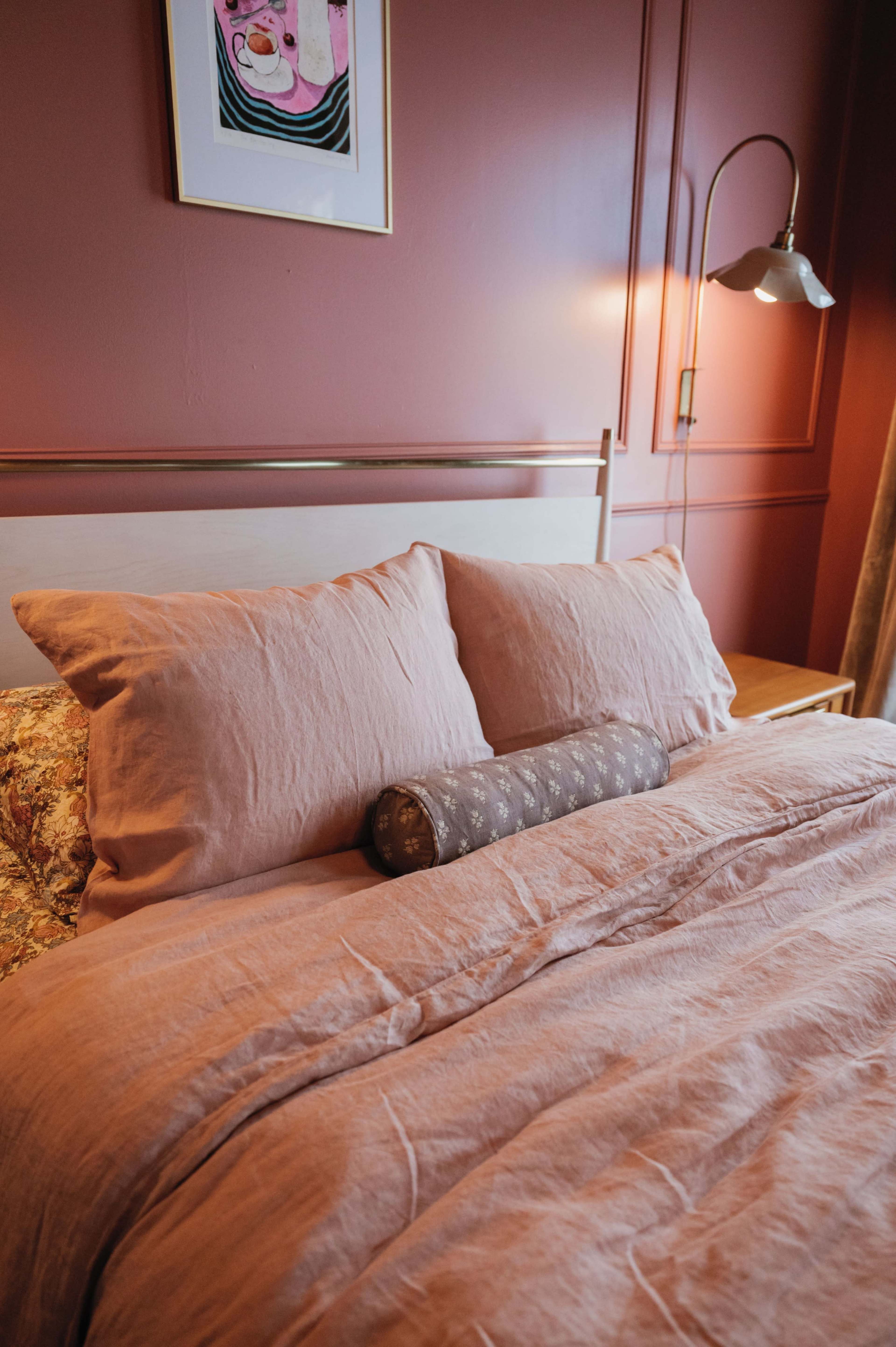 The image shows a neatly made bed with large, soft pillows and a decorative bolster pillow against a wall painted in a warm, muted color.