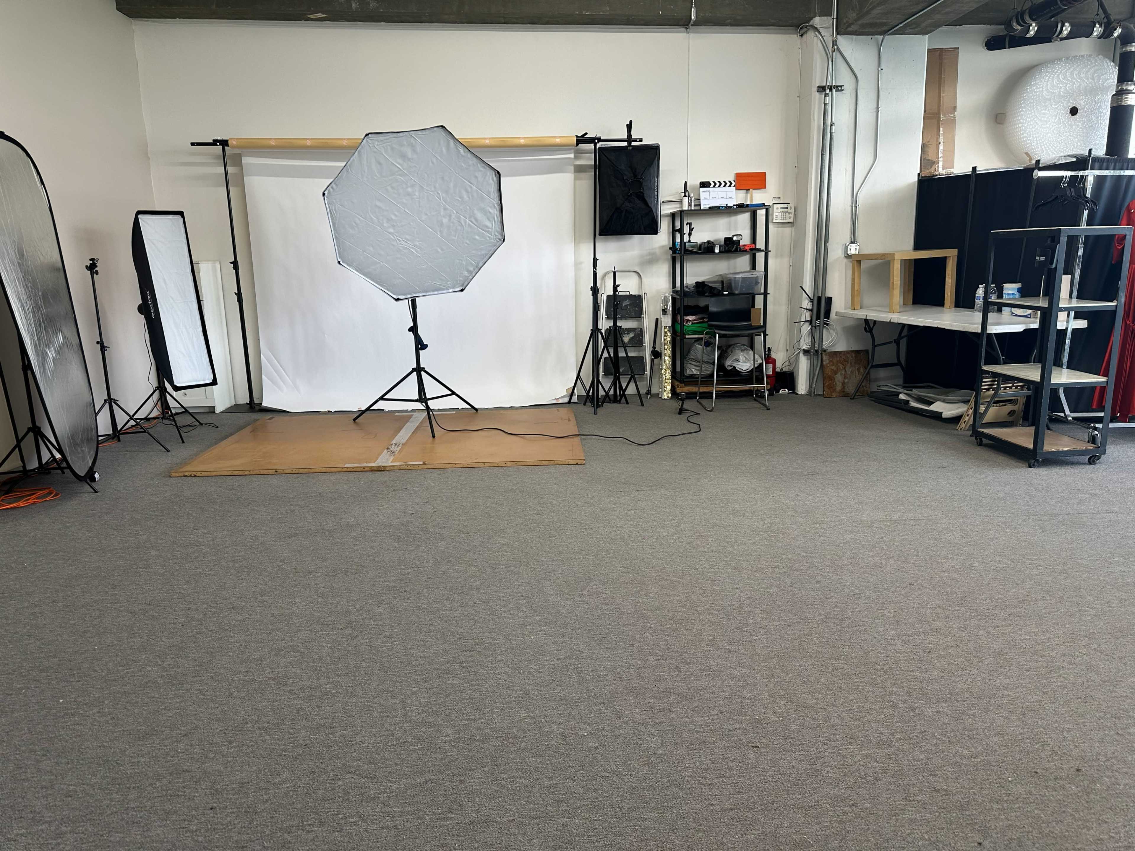 The image shows an empty photography studio with a white backdrop, an octagonal softbox light, and various equipment arranged on shelves.