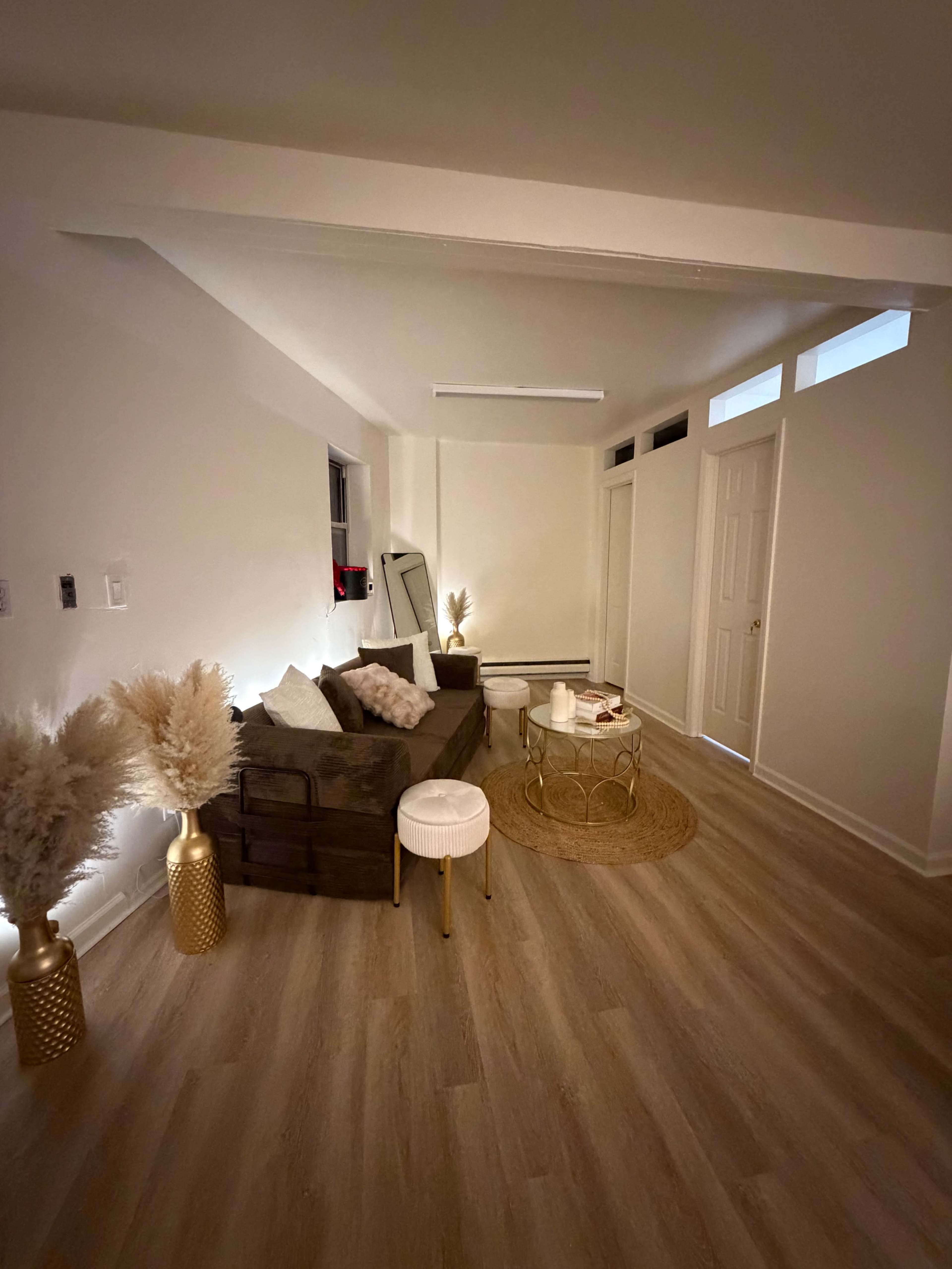 The scene features a sparsely furnished living room with a brown couch, a round golden coffee table, and decorative plants in gold vases, all set against light-colored walls and flooring.