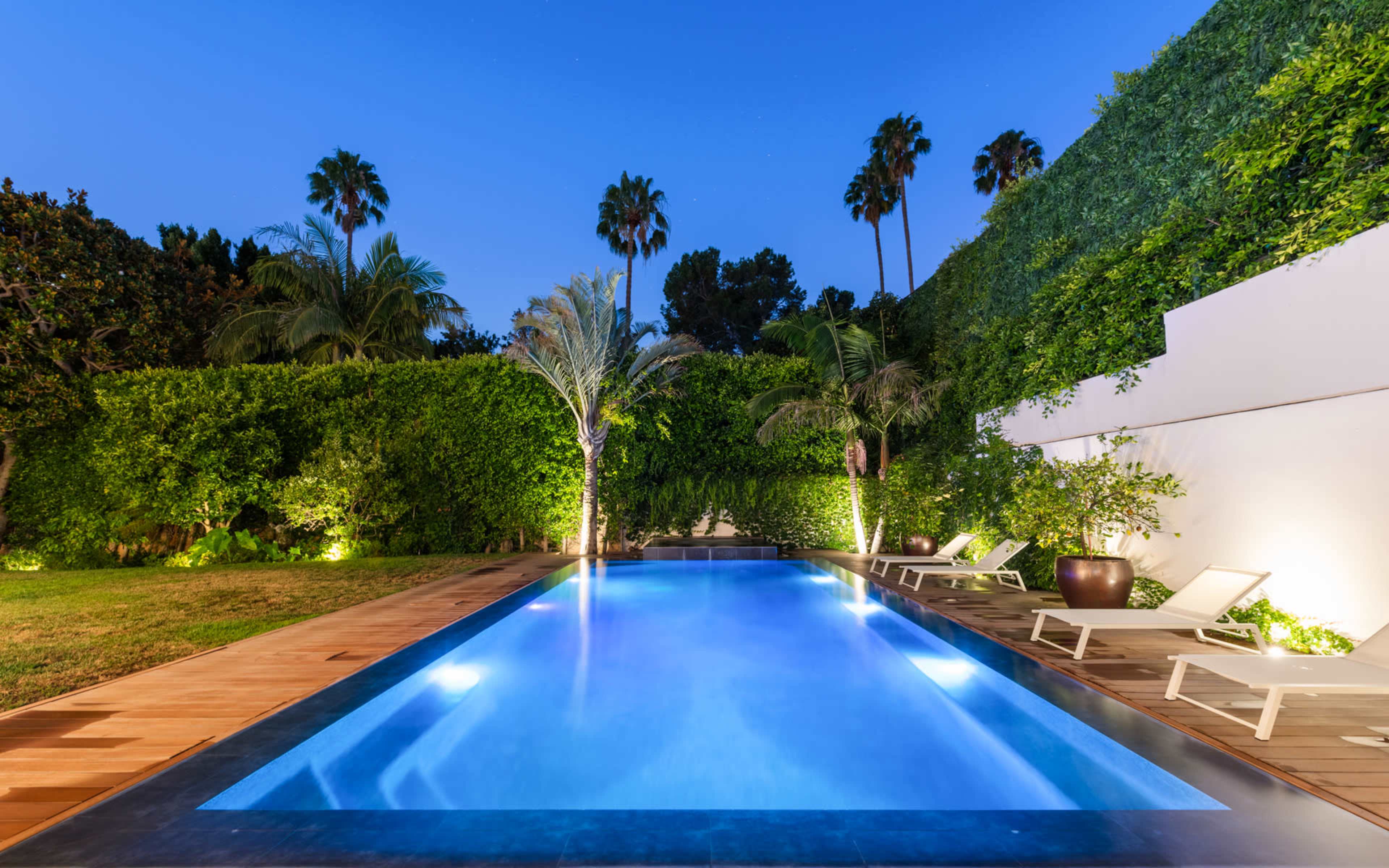 The image shows a modern swimming pool surrounded by lush greenery and lounge chairs under a twilight sky.