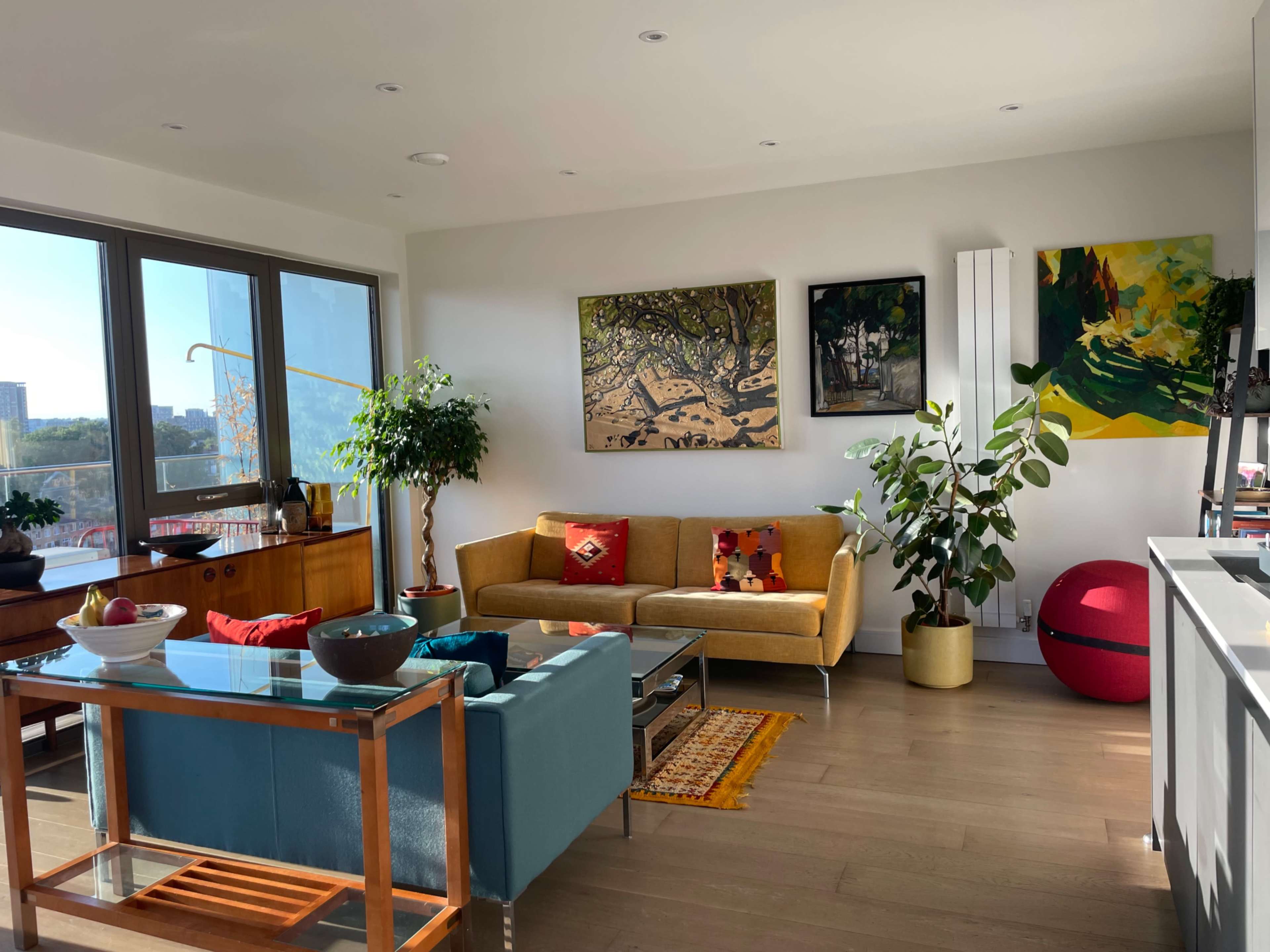 A bright, modern living room with a yellow sofa, a glass coffee table, potted plants, and large windows offering a view of the city.