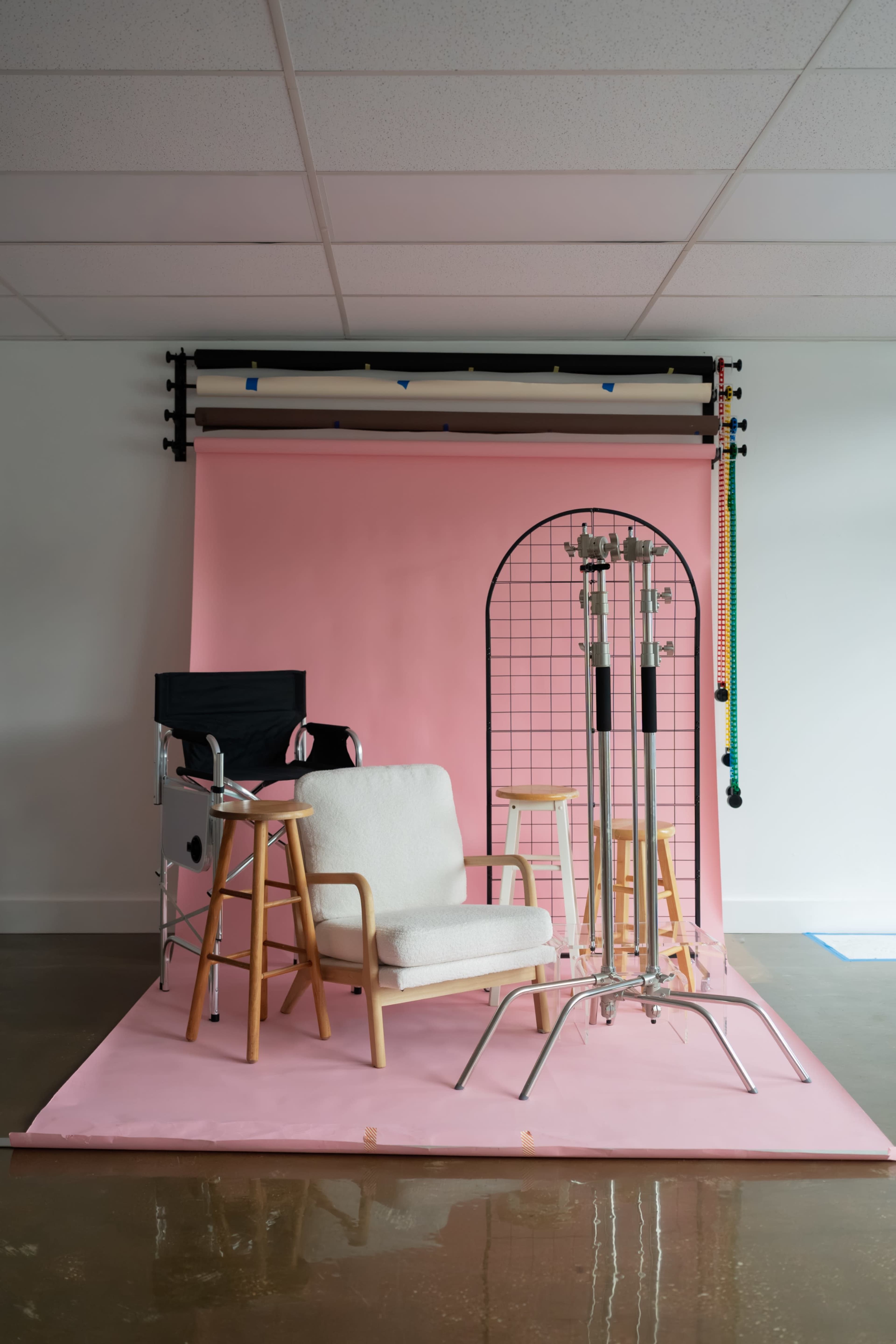 The image shows a photography studio setup featuring a light pink backdrop, a white upholstered chair, two wooden stools, and a metal stand for studio equipment.