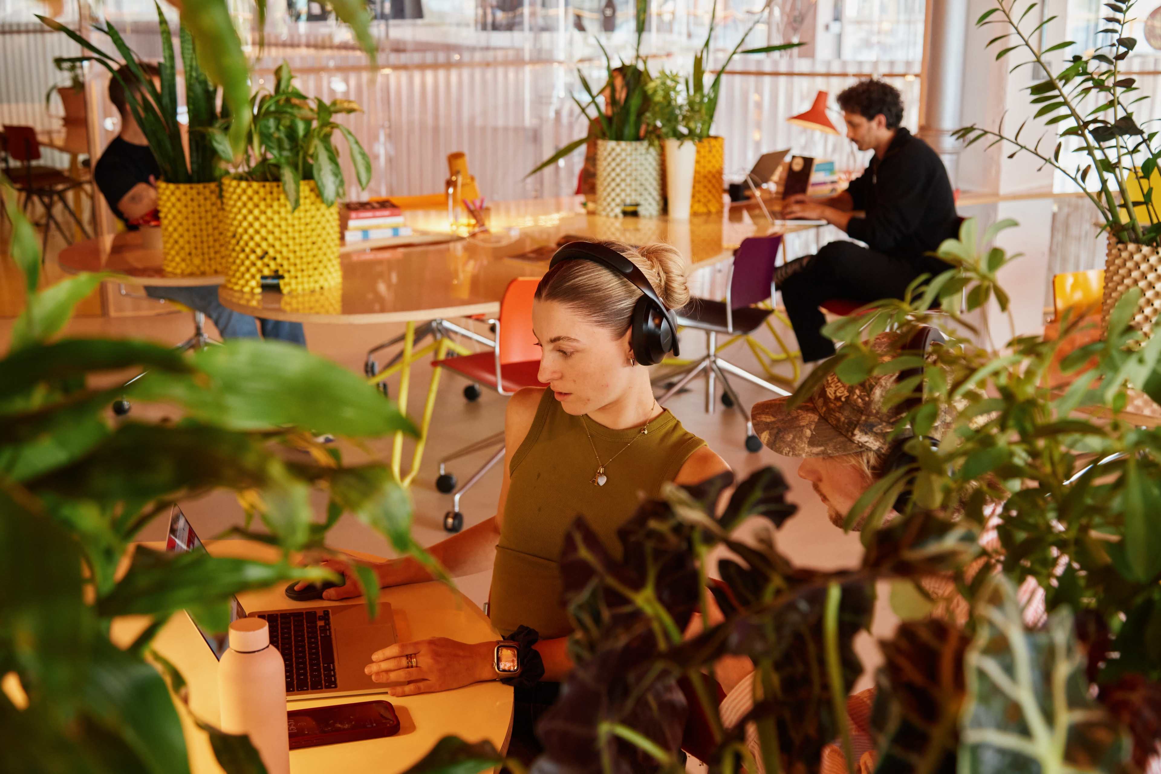 A woman wearing headphones works on a laptop at a brightly lit office space filled with plants, while two other individuals are engaged in different activities in the background.