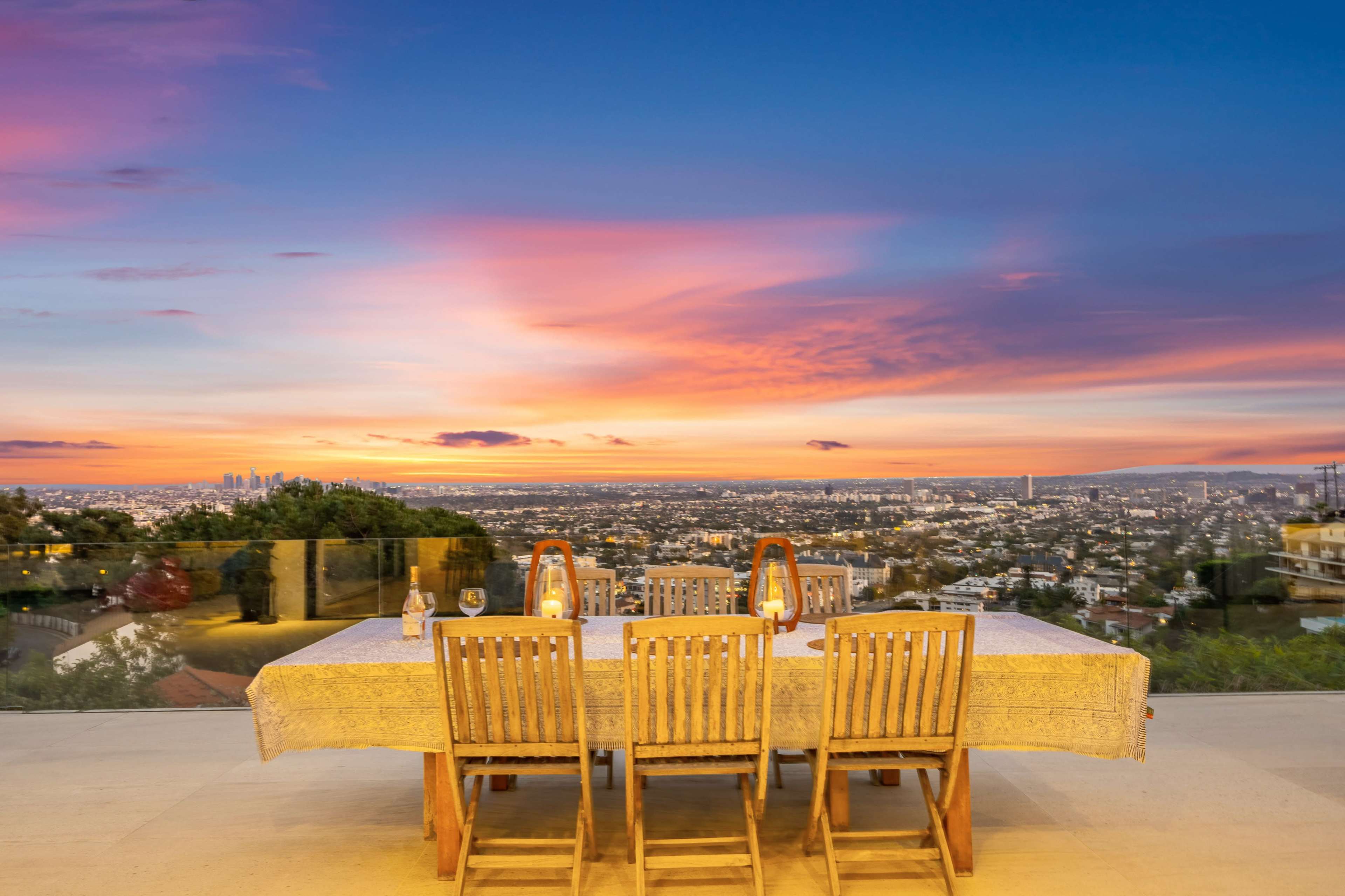 A dining table with wooden chairs set against a backdrop of a vibrant sunset over a city skyline.
