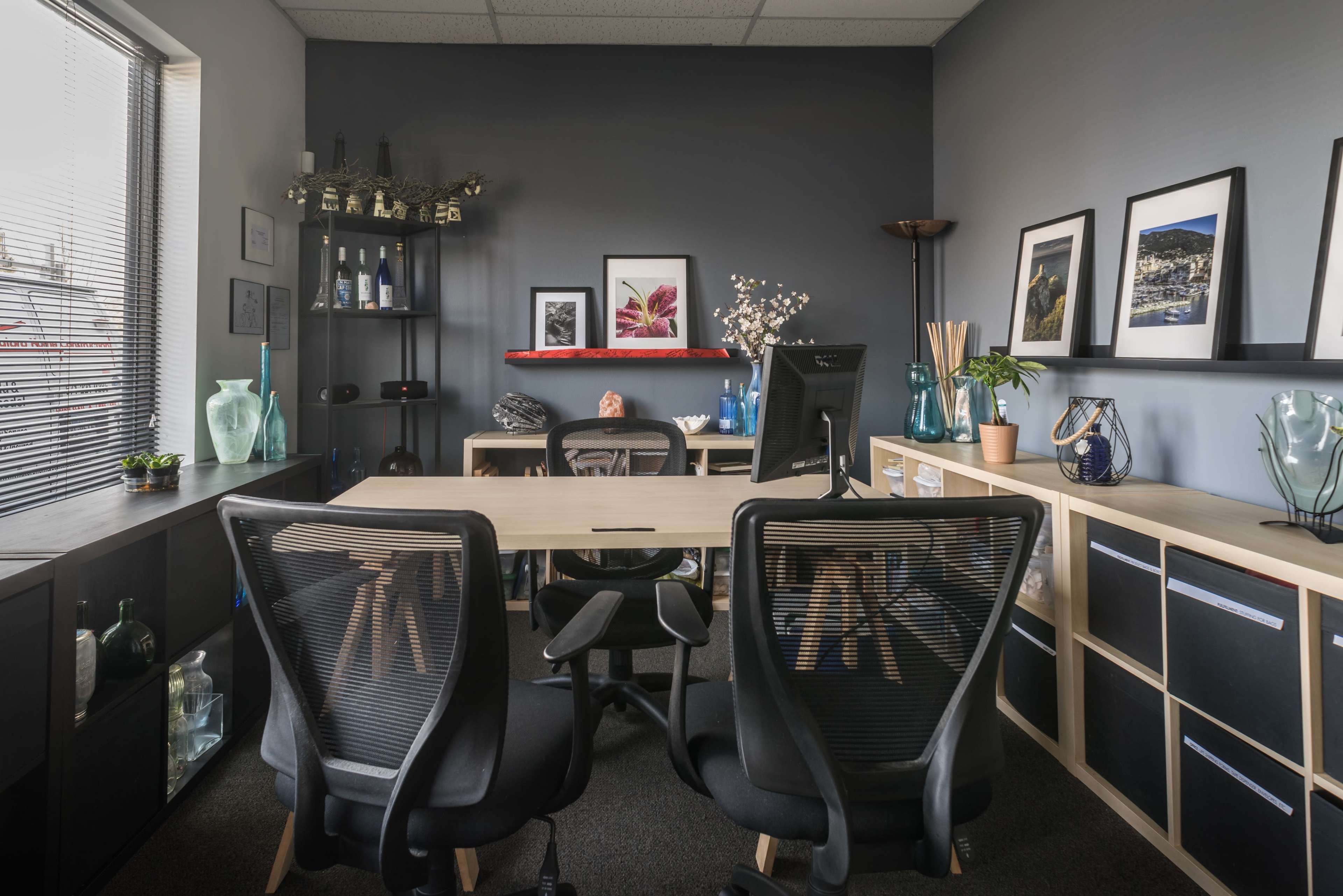 A modern office space features a desk with black chairs, shelves displaying decorative items, and framed artwork on the walls.