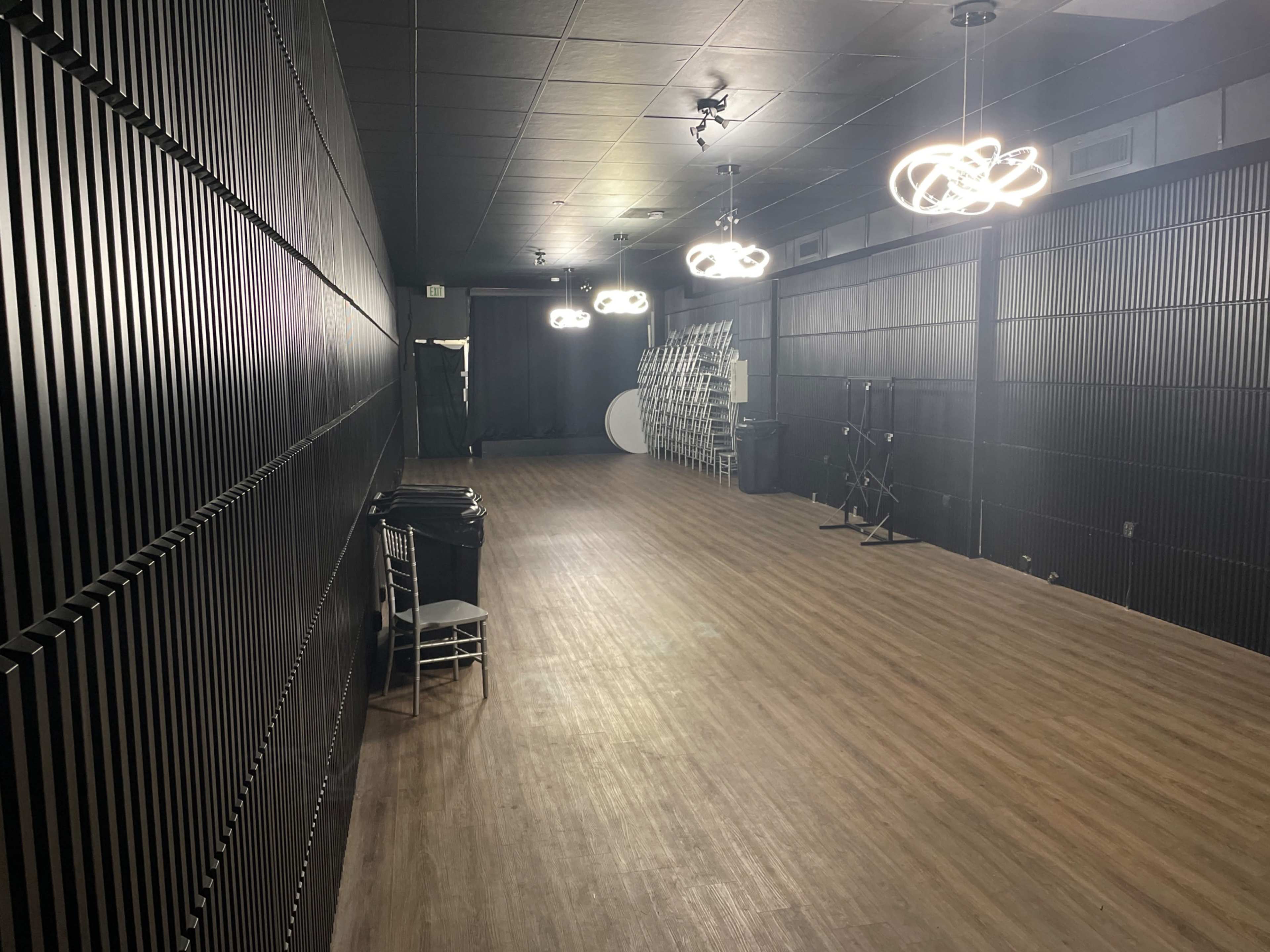 A long, narrow room with dark walls, modern light fixtures, and wooden flooring, featuring various furniture and equipment along one side.