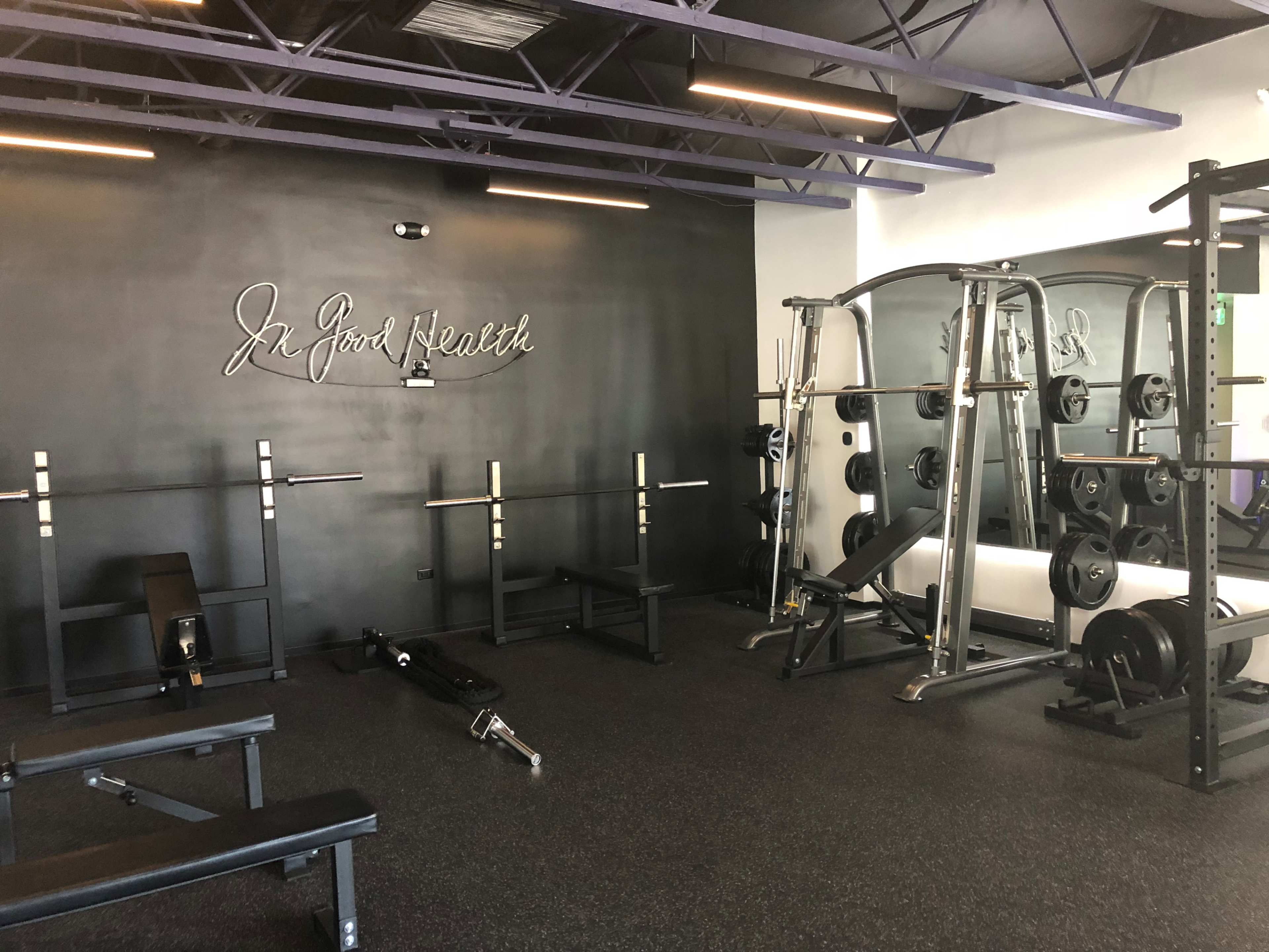 A gym interior featuring weightlifting equipment, benches, and a wall sign that reads "In Good Health."