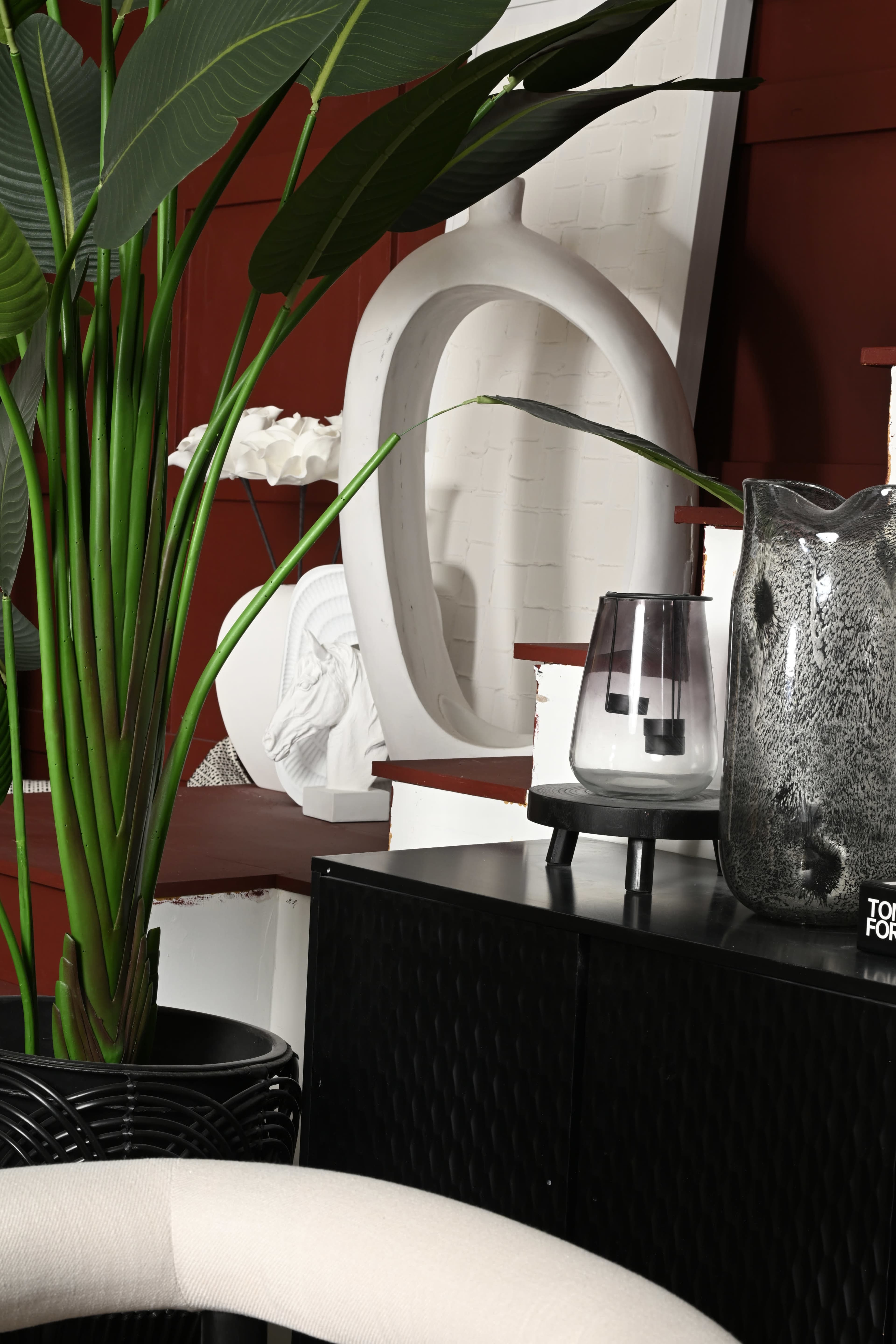 The image features a modern interior scene with tall green plants, a matte black cabinet, a decorative glass vase, and an abstract white sculpture against a red wall.