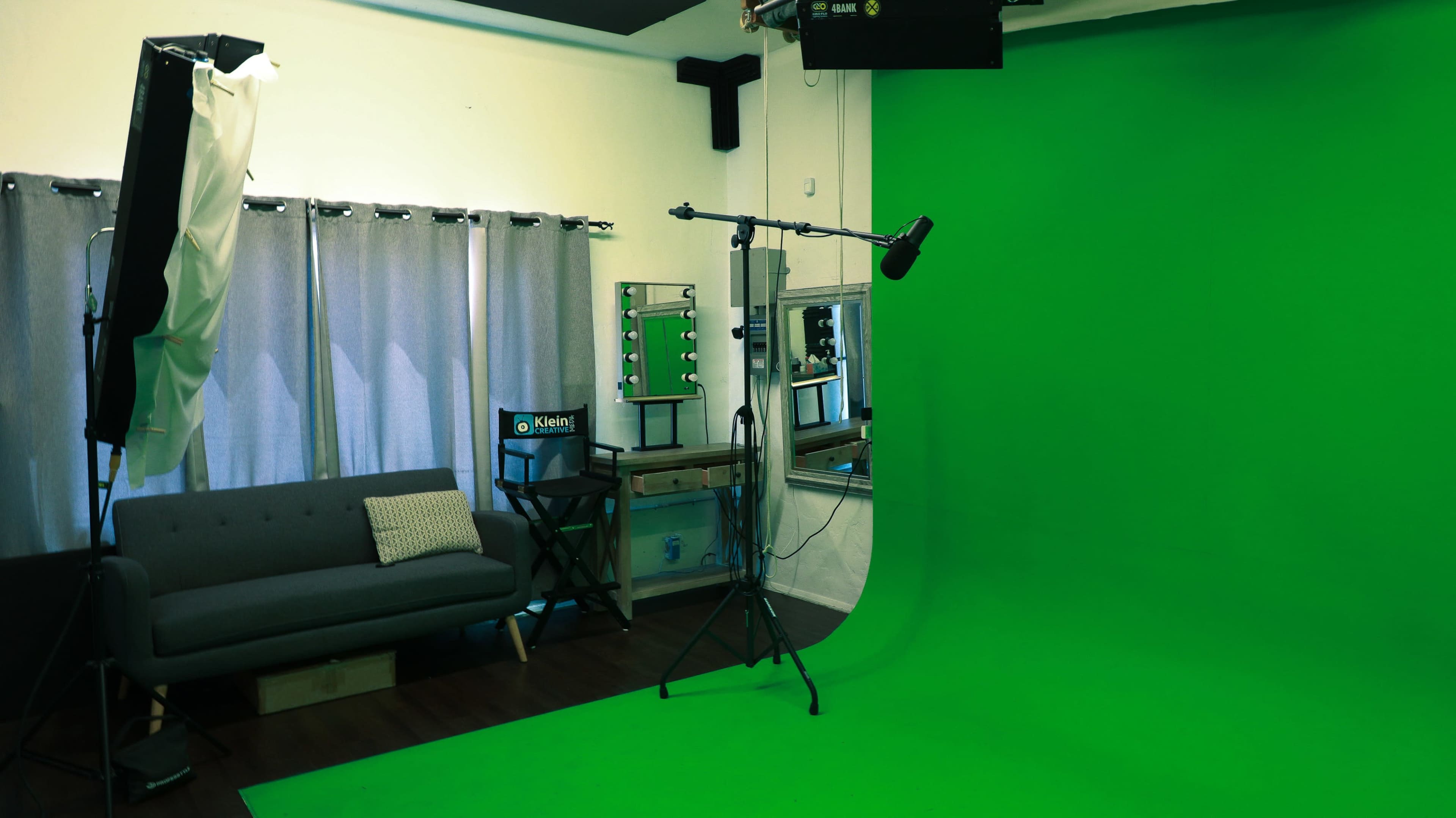 The image shows a green screen studio with a gray sofa, curtains, a mirror, and lighting equipment.