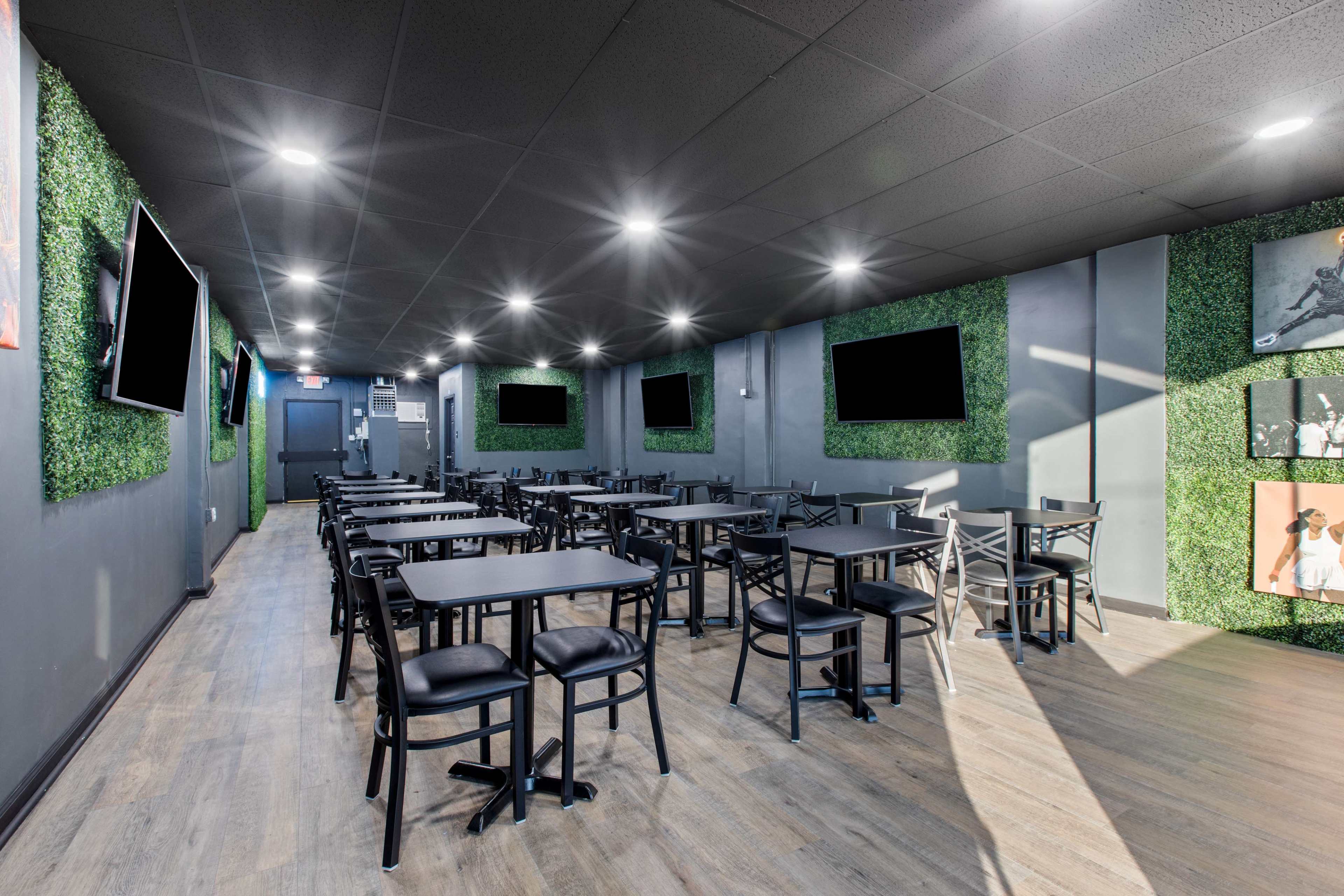 A modern restaurant interior with black tables and chairs, green wall decorations, and several wall-mounted screens.