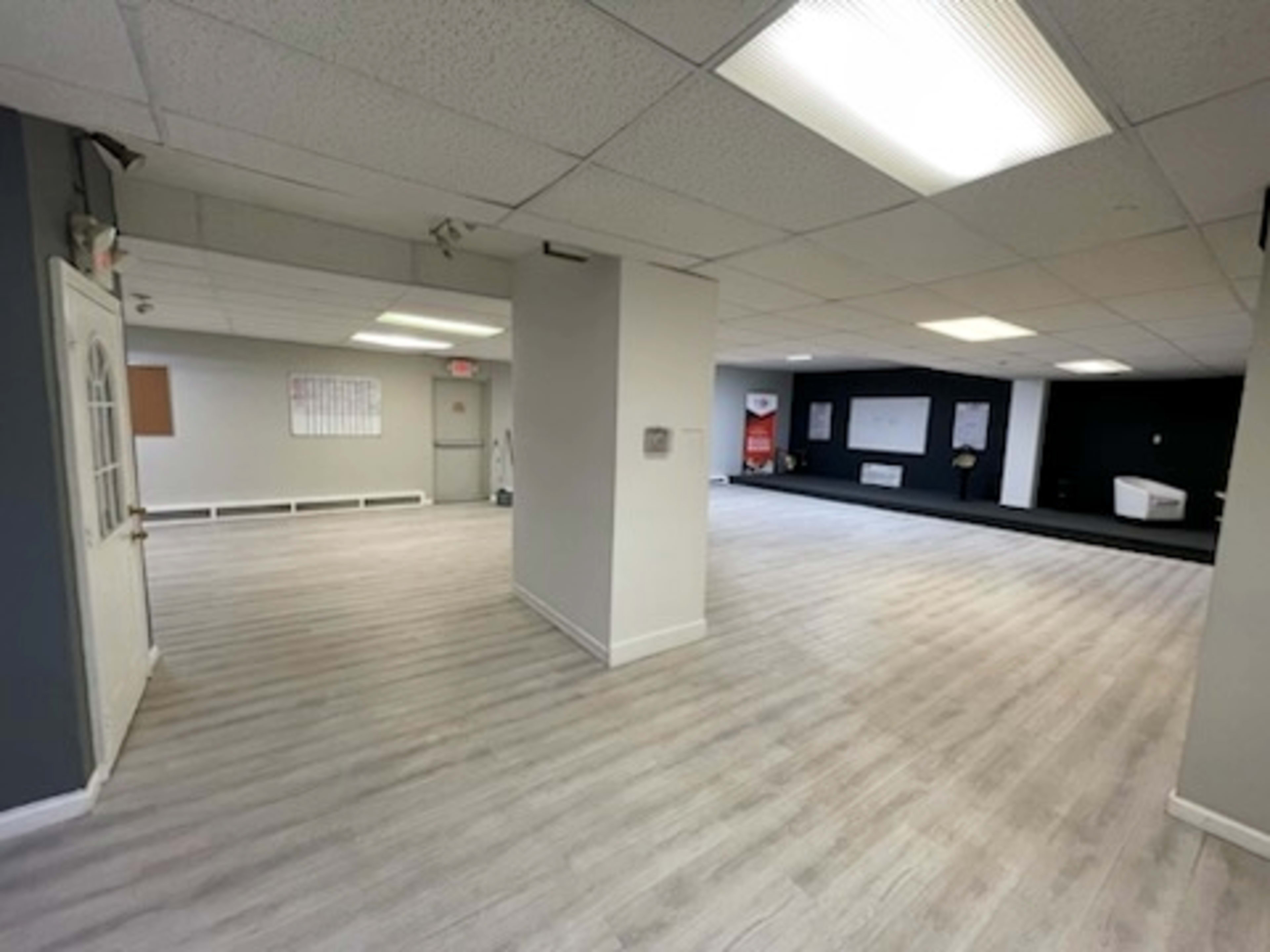 The image shows a spacious interior room with light-colored wooden flooring, white walls, and several overhead fluorescent lights.