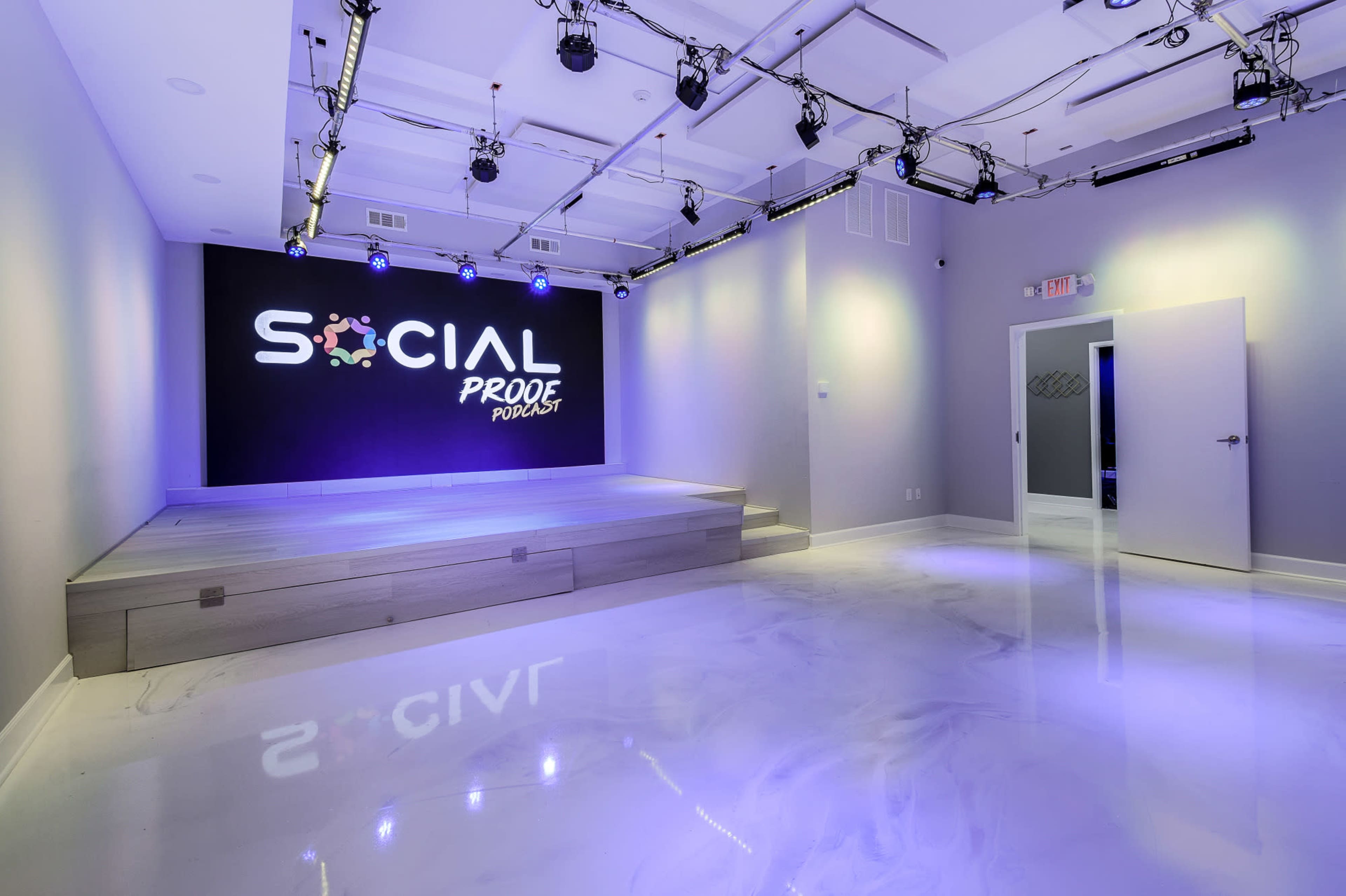 The image shows a modern event space with a raised platform, illuminated by soft purple lighting, featuring a wall that reads "SOCIAL PROOF PURSUIT."