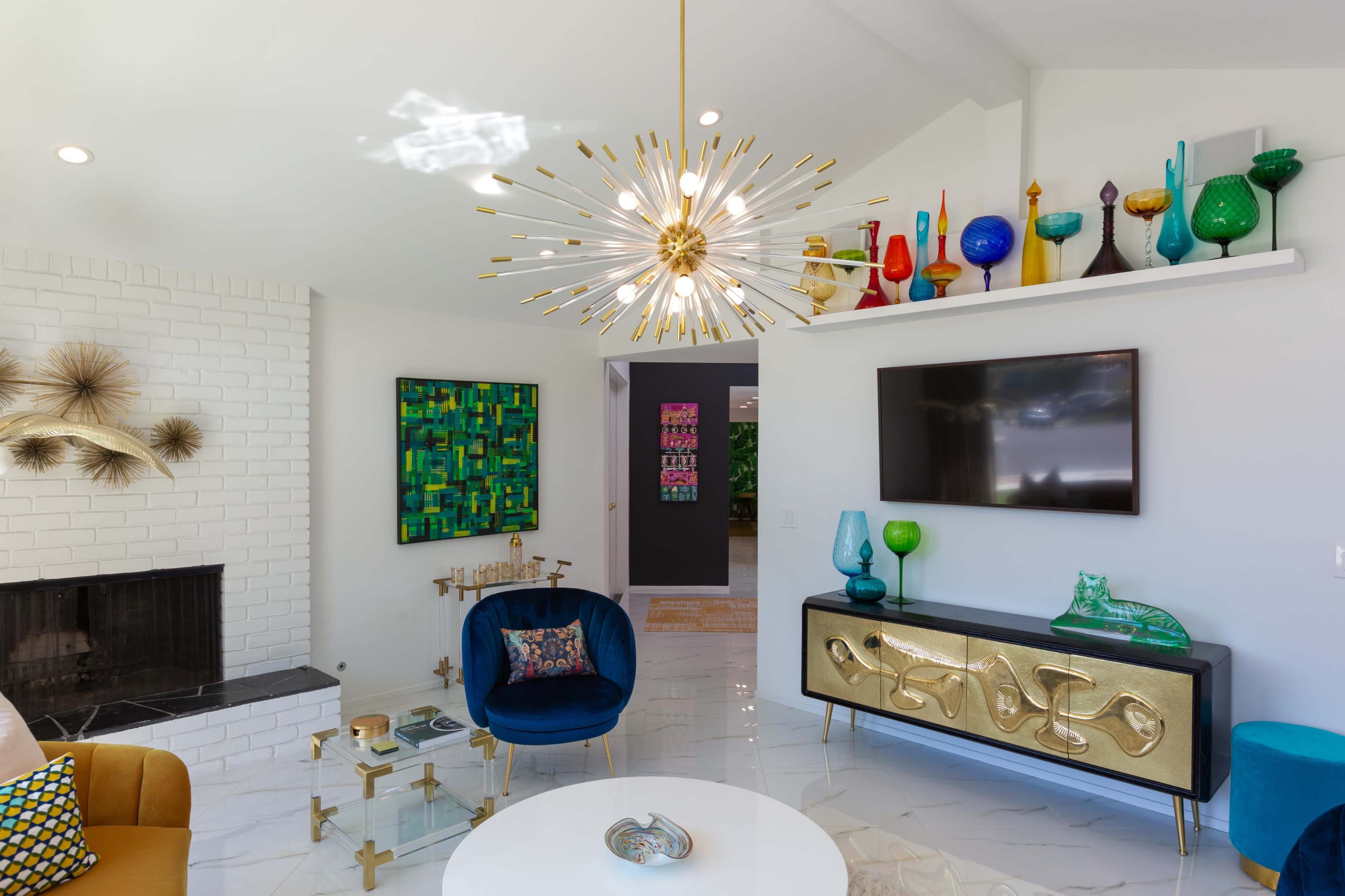 A modern living room features a gold starburst chandelier, colorful glass vases on a shelf, and a blue velvet chair.