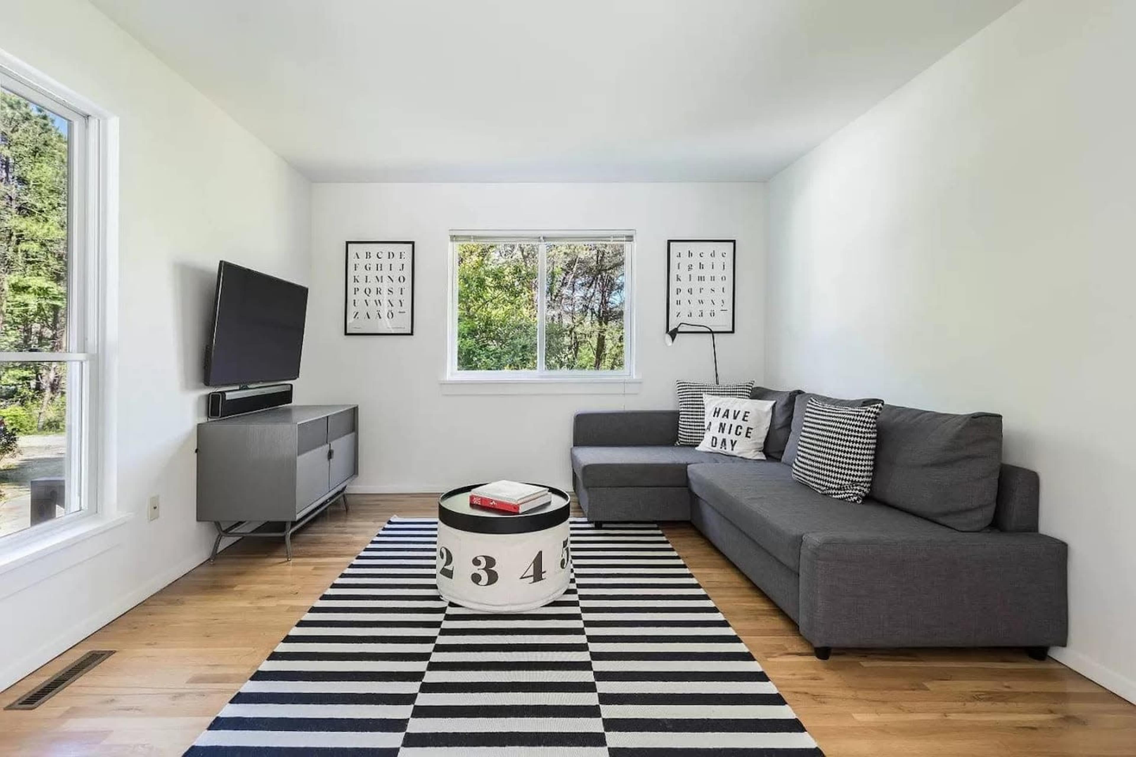 A modern living room featuring a gray sectional sofa, a black and white striped rug, a coffee table with bold numbers, and a television mounted on the wall.