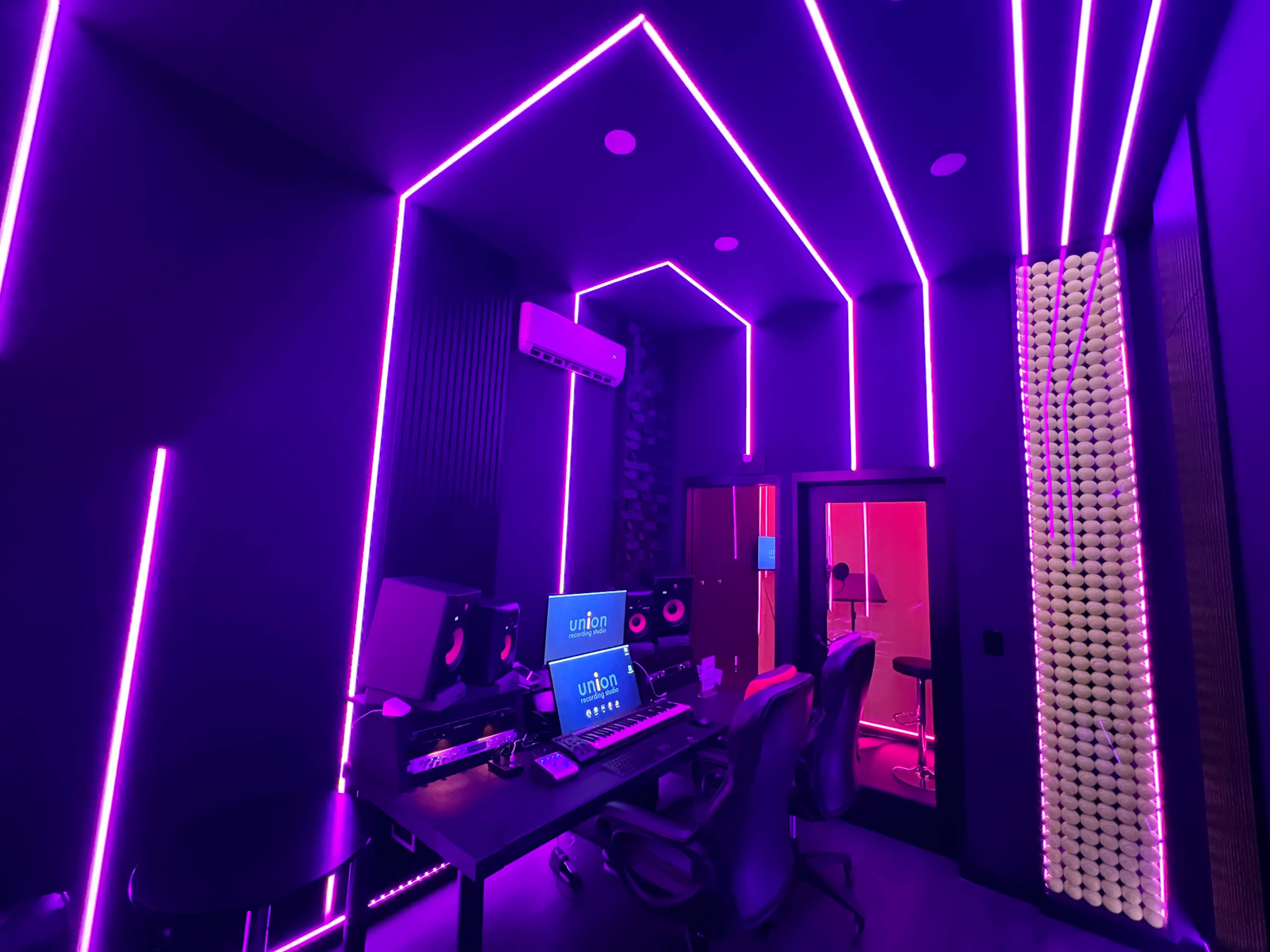 The image shows a music studio with purple LED lighting, featuring a central workstation and a separate recording room.