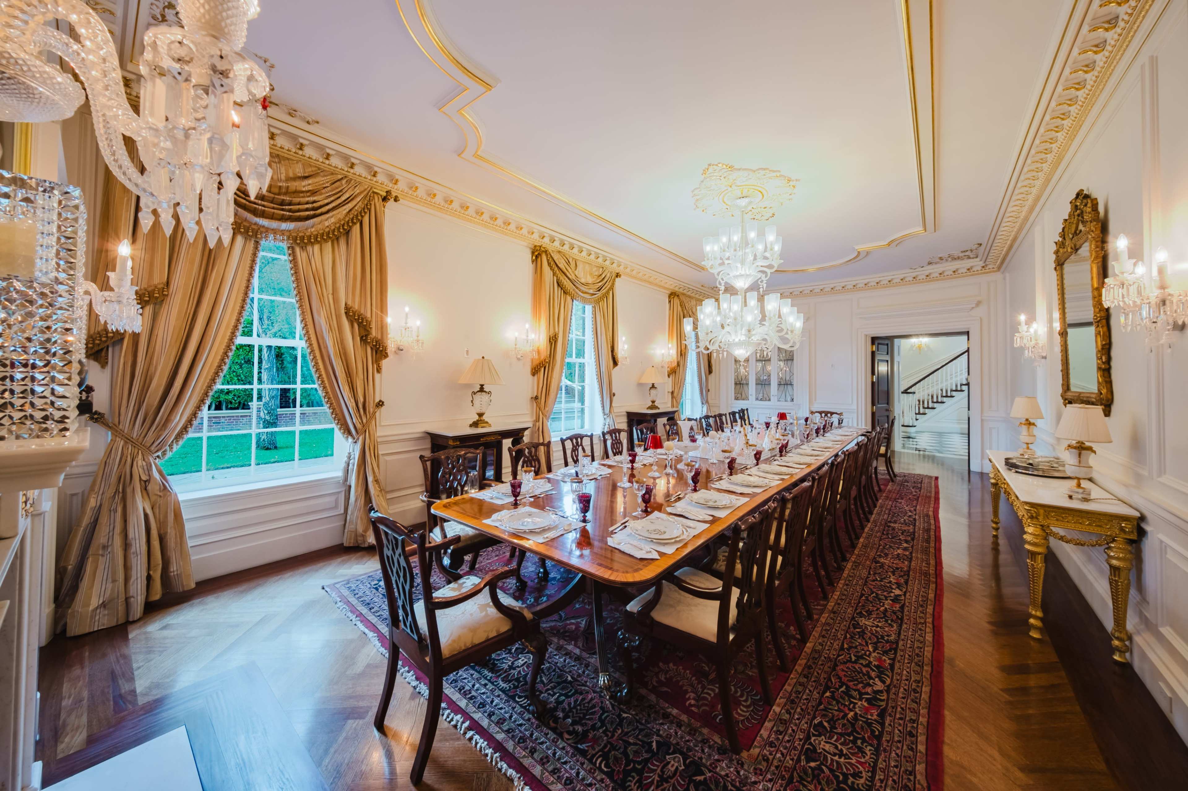The image shows an elegantly appointed dining room featuring a long wooden table set for a meal, surrounded by ornate chairs and adorned with crystal chandeliers and luxurious drapery.