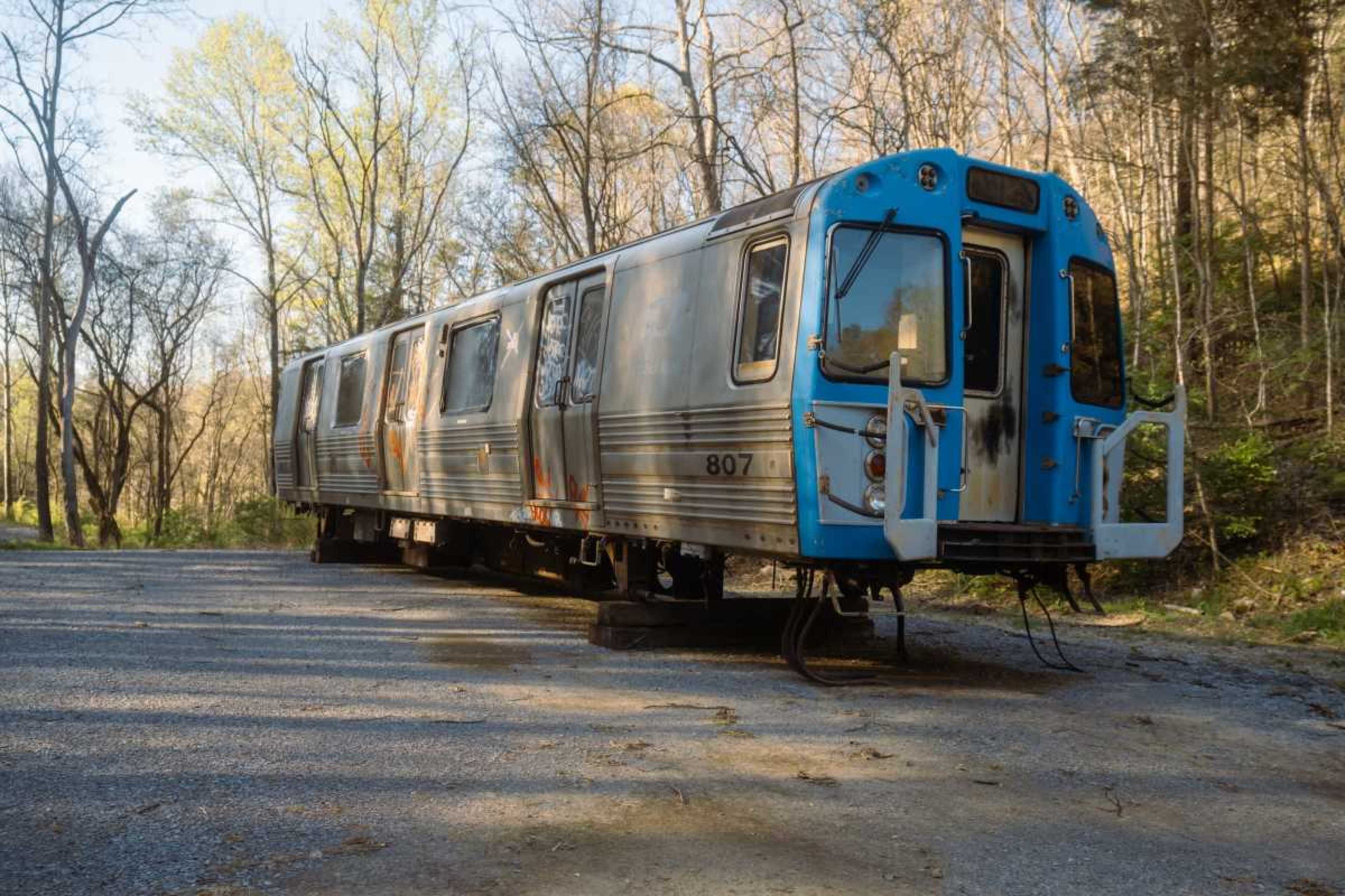 An abandoned subway train car, with rusted metal and a blue front, is parked on a gravel path surrounded by trees.