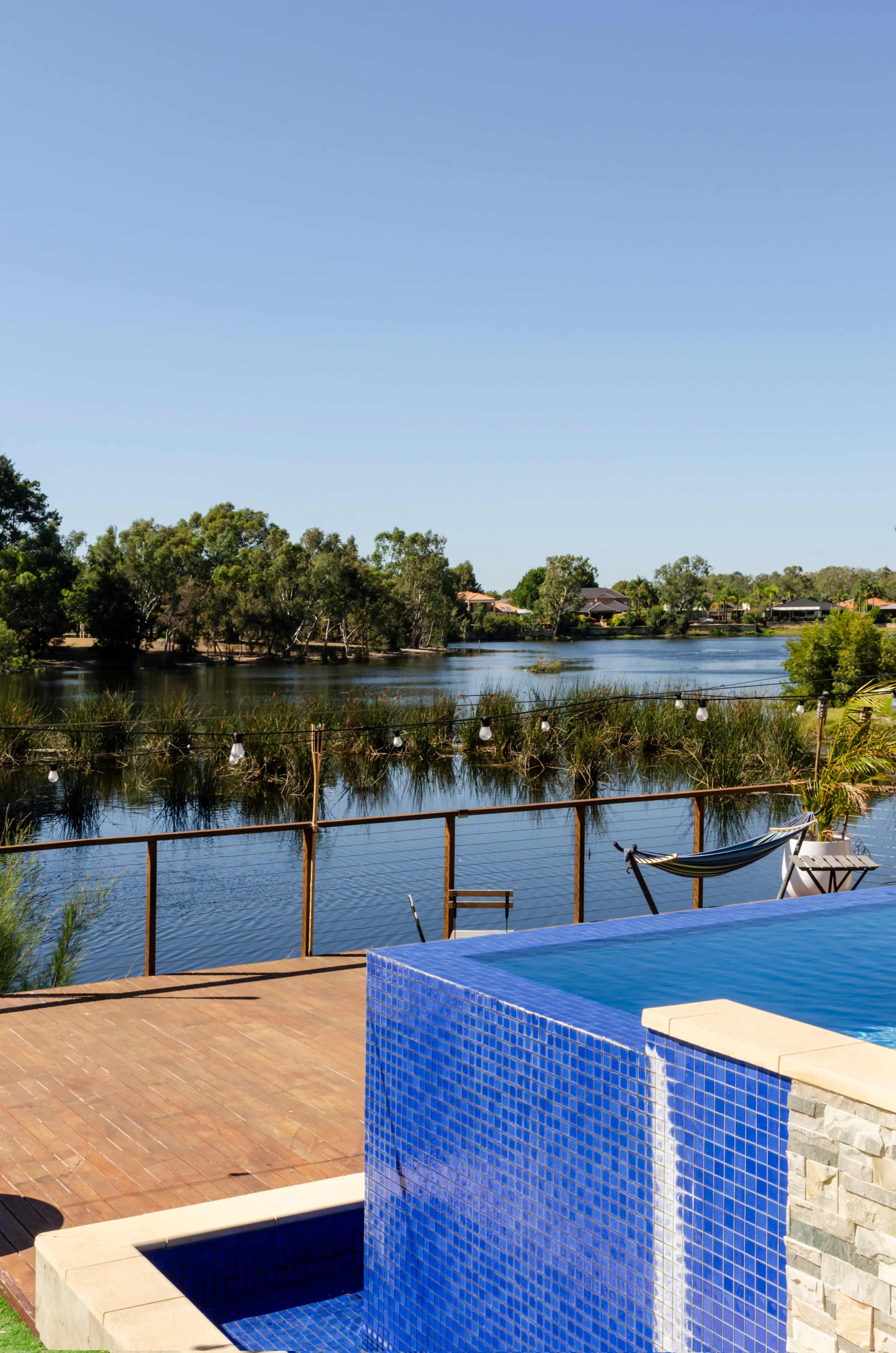 A blue-tiled infinity pool overlooks a calm river, with greenery and houses visible across the water.
