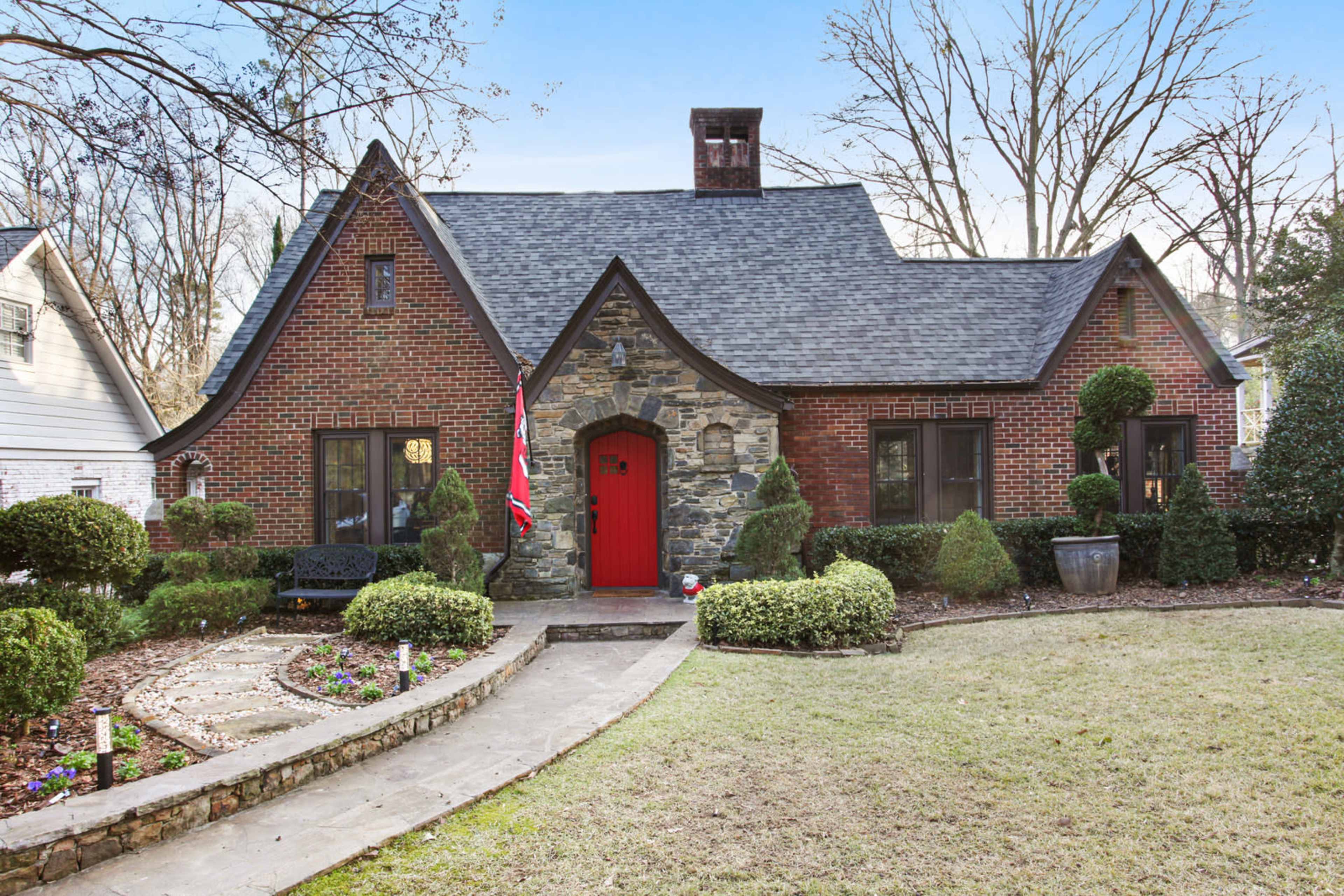 A stone and brick house features a red door, surrounded by manicured bushes and a gravel pathway.