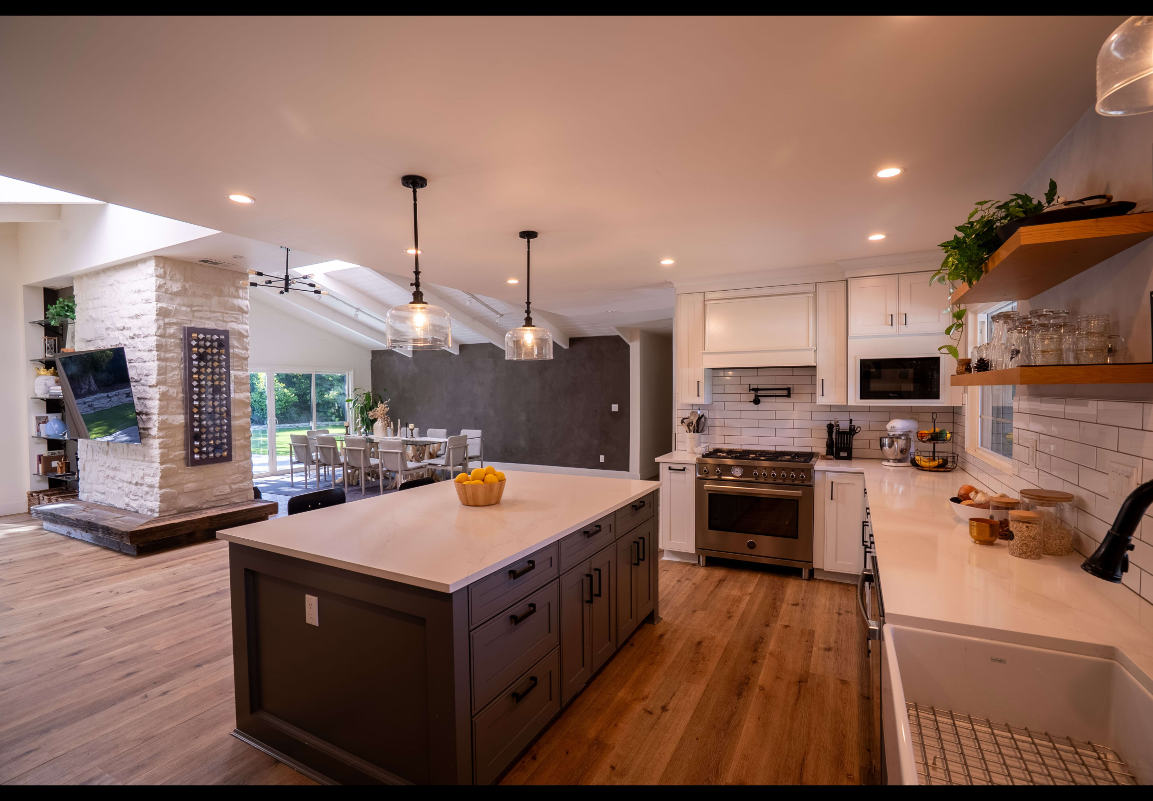 The image shows a modern kitchen with a large island, stainless steel appliances, and a dining area visible in the background, featuring a mix of white and dark cabinetry.
