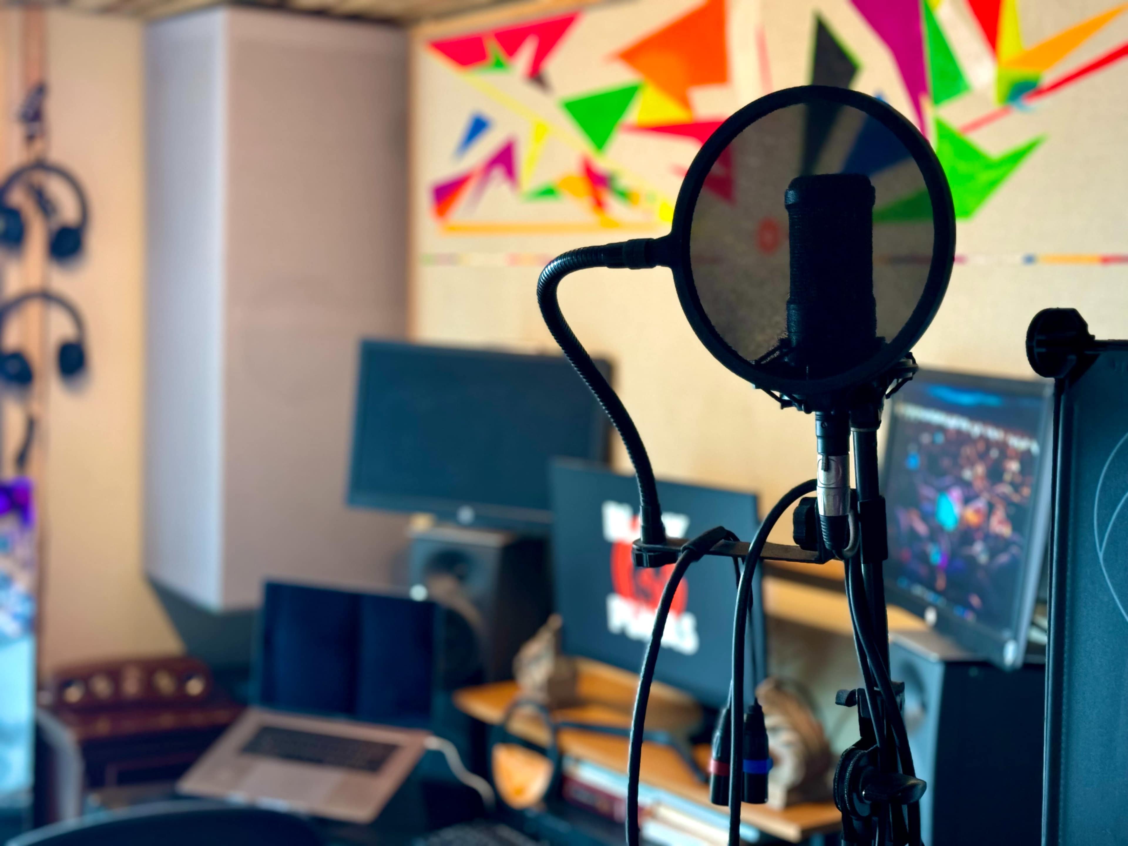 A microphone stand with a pop filter is positioned in a home recording studio filled with computers and colorful wall art.