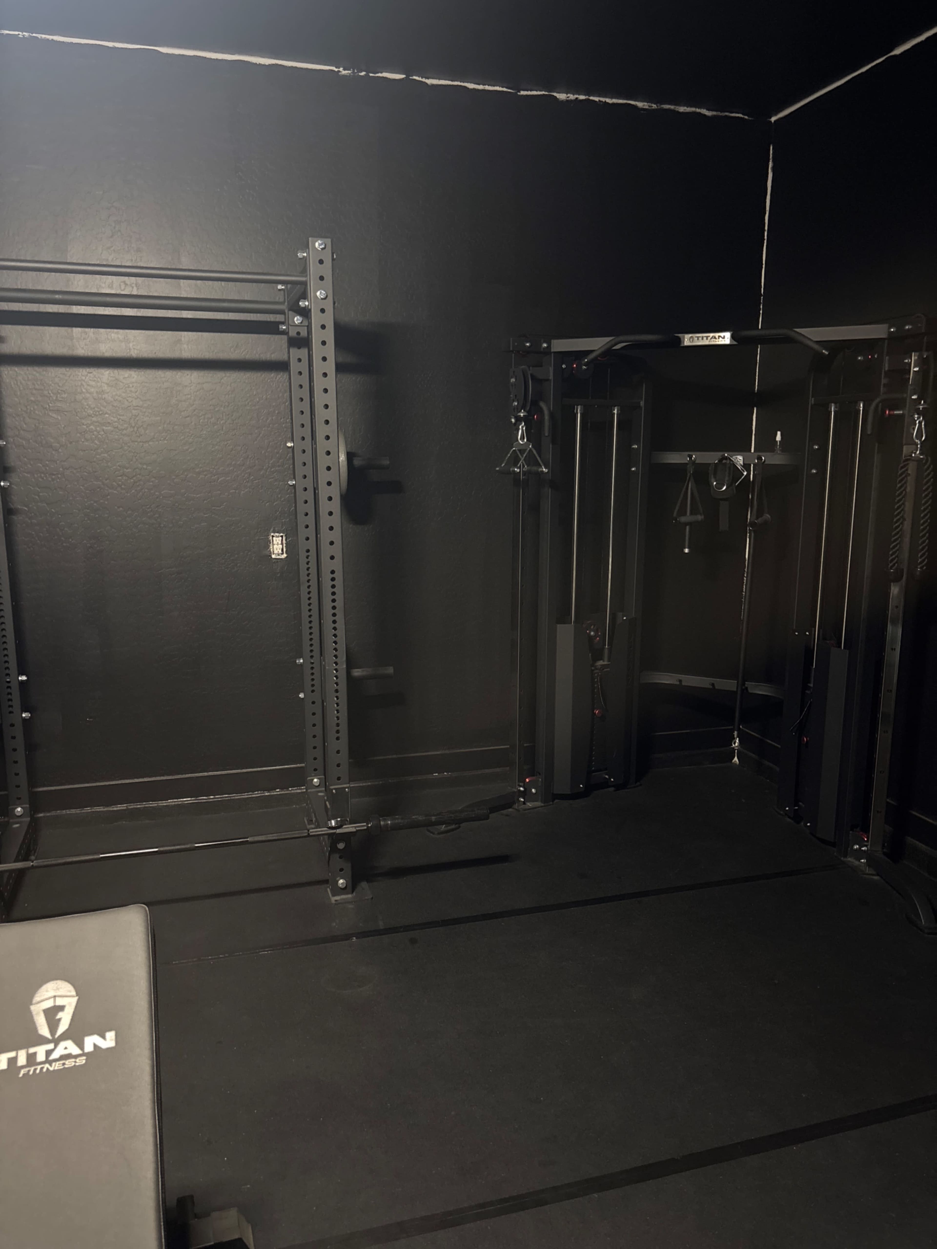 The image shows a sparse gym space featuring a bench, cable machines, and a rack system against black walls.