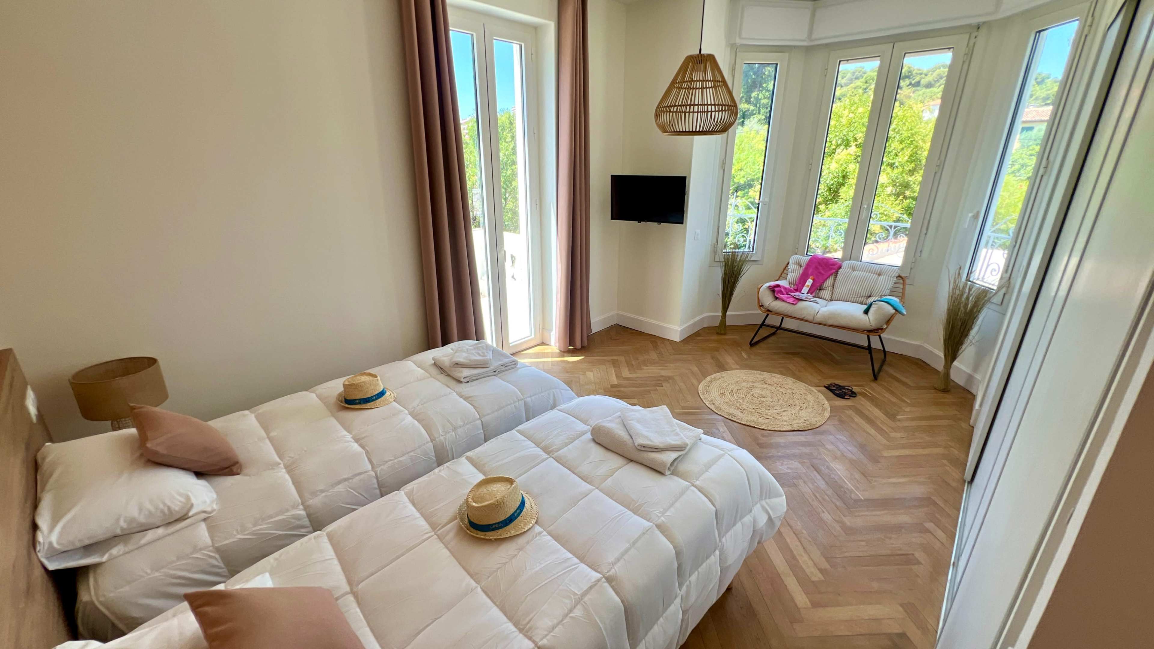 The image shows a bright, airy bedroom with two twin beds, a television mounted on the wall, and a cozy seating area by a large window.
