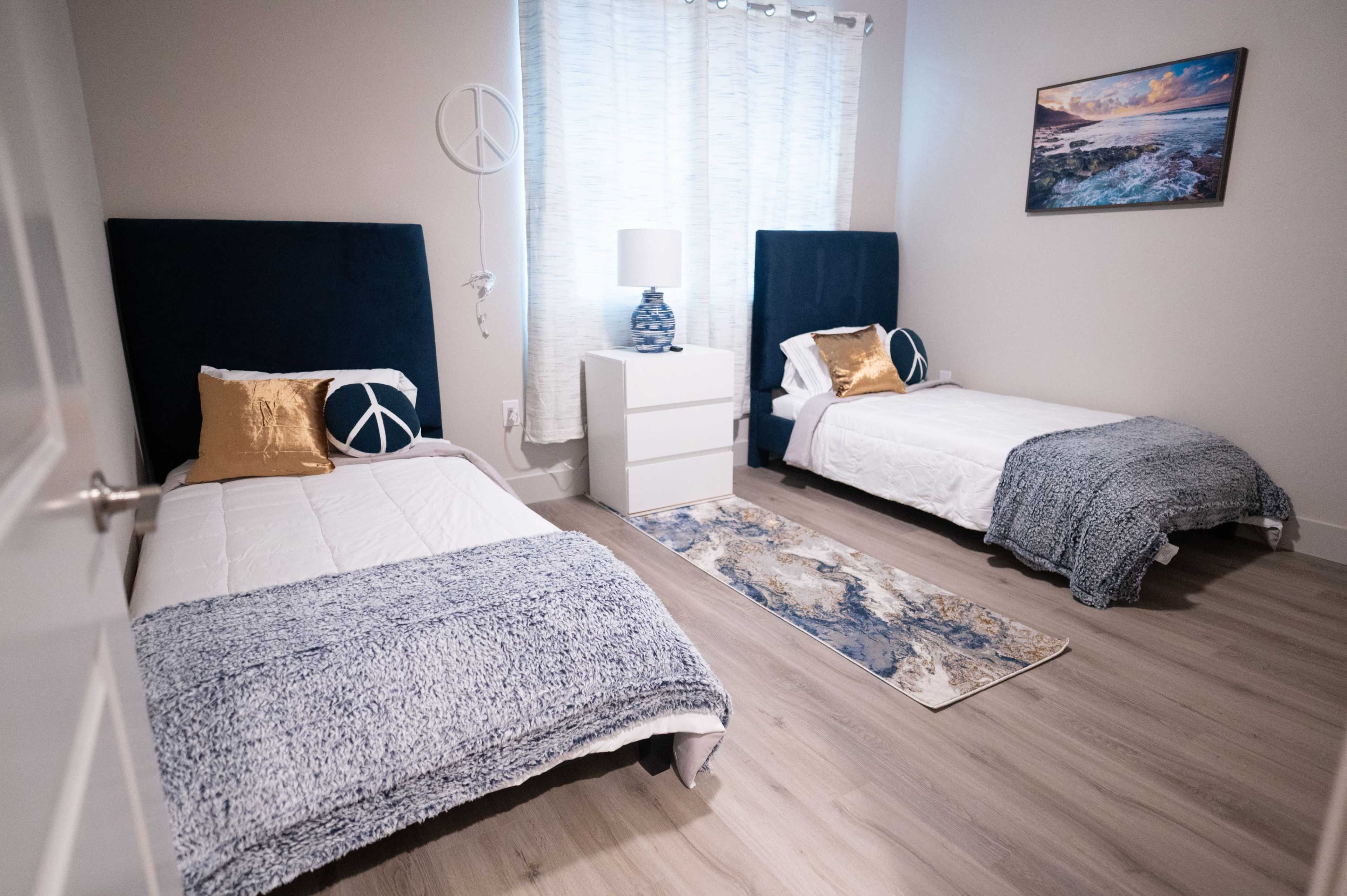 The image shows a spacious bedroom with two twin beds, a white dresser, and a window with sheer curtains, featuring a peaceful blue and white color scheme.
