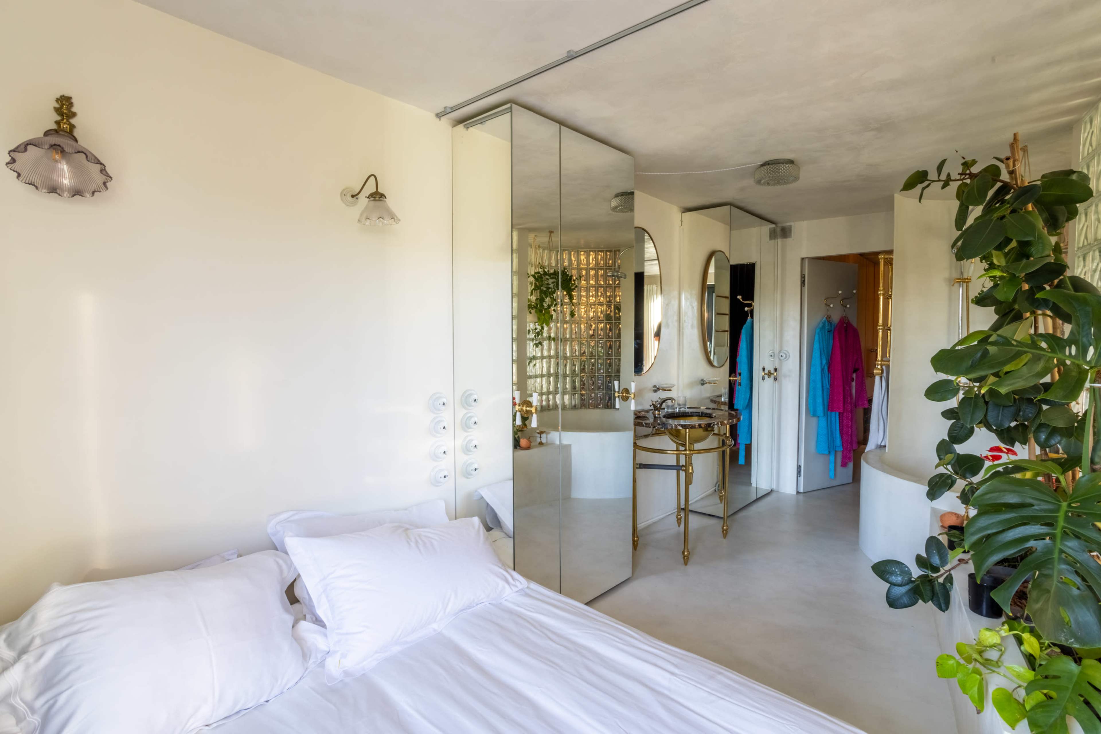 The image shows a minimalist bedroom with a white bed, mirrored closet, and plants, leading into a bright bathroom area.