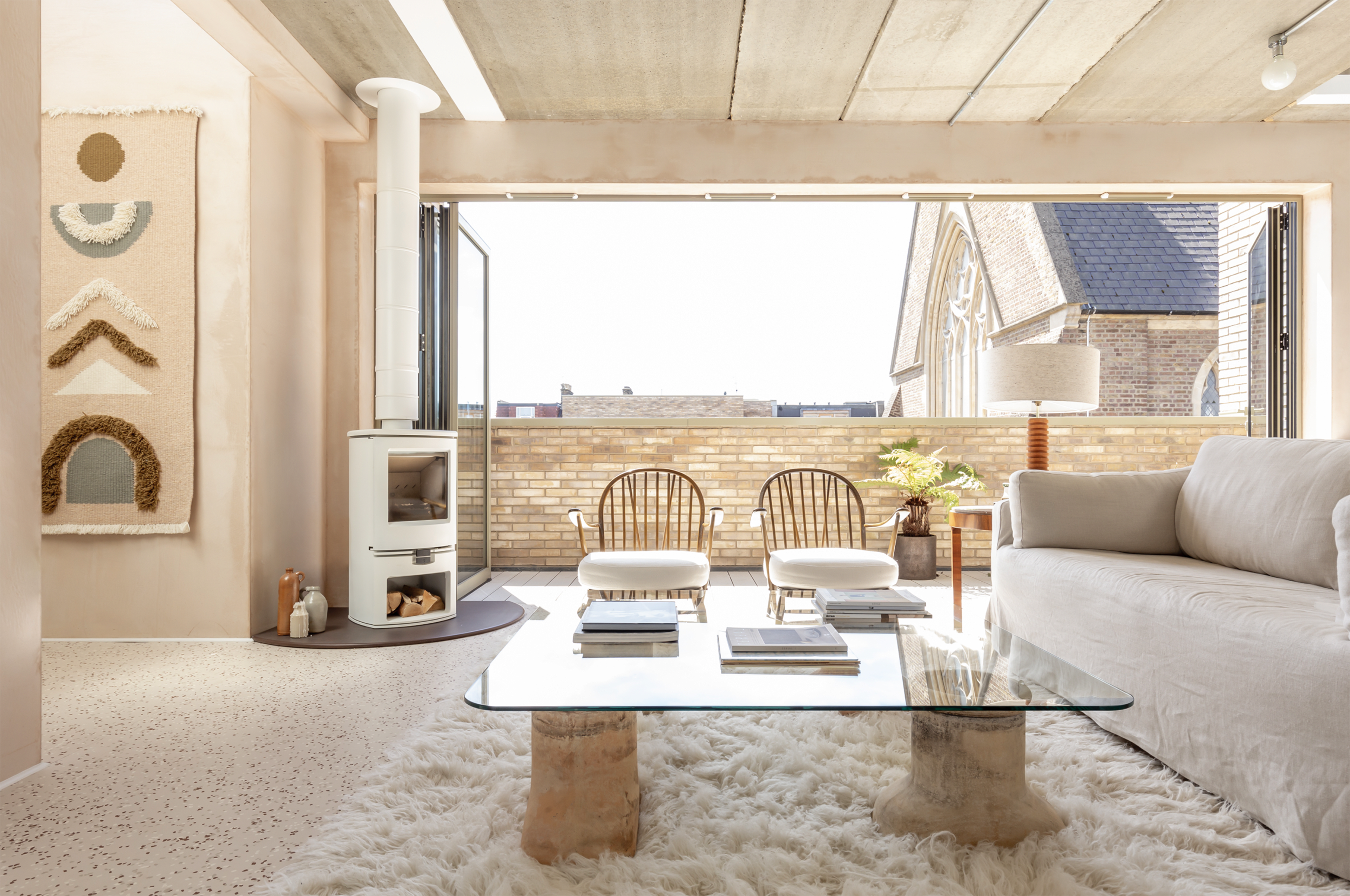 A modern living room features a white sofa, two wicker chairs, a glass coffee table, and large windows that open to a view of rooftops.