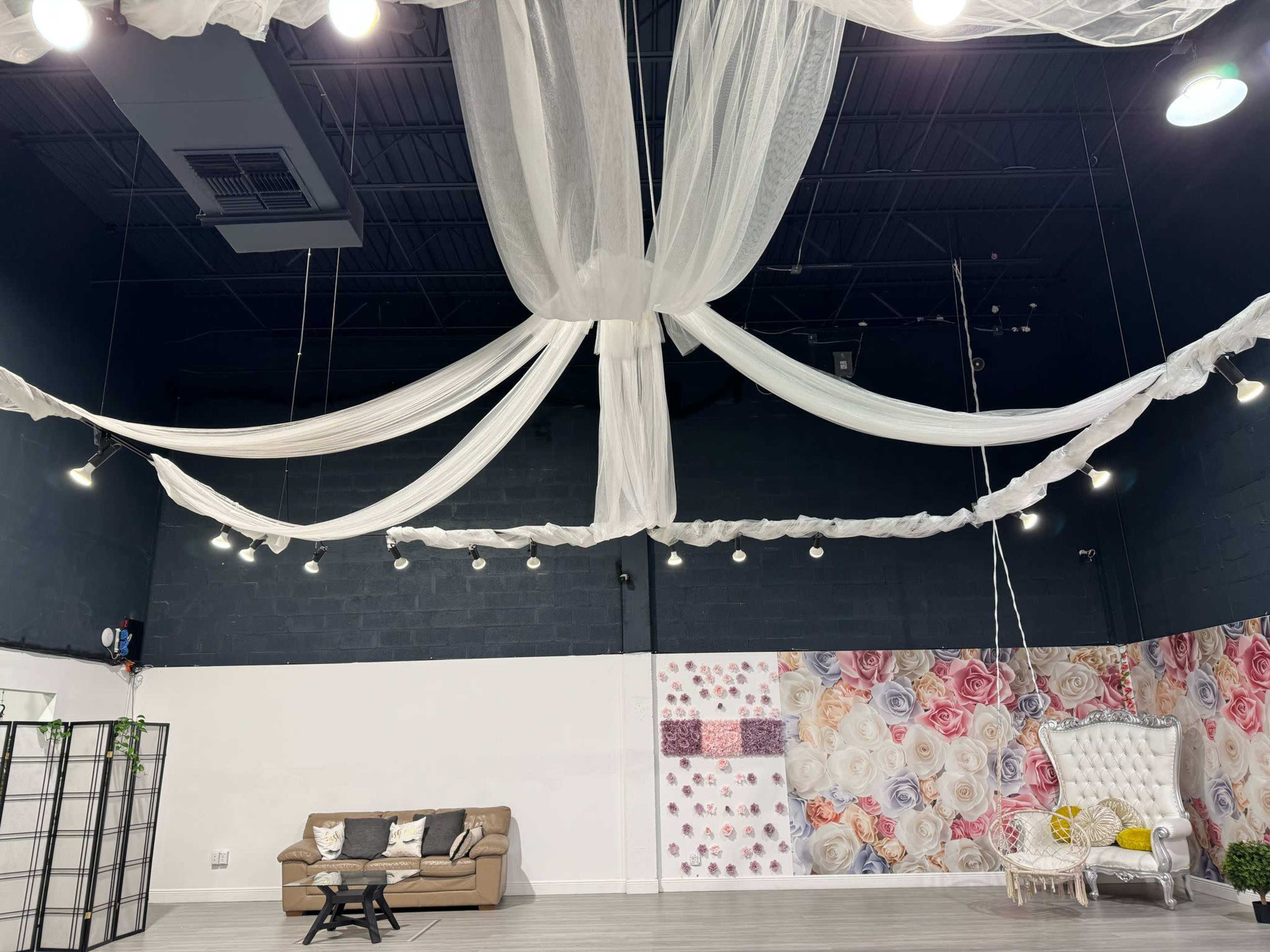 The image shows an event space with draped white fabric across the ceiling, a beige sofa in the foreground, and a floral-patterned wall sectioned off to the right.